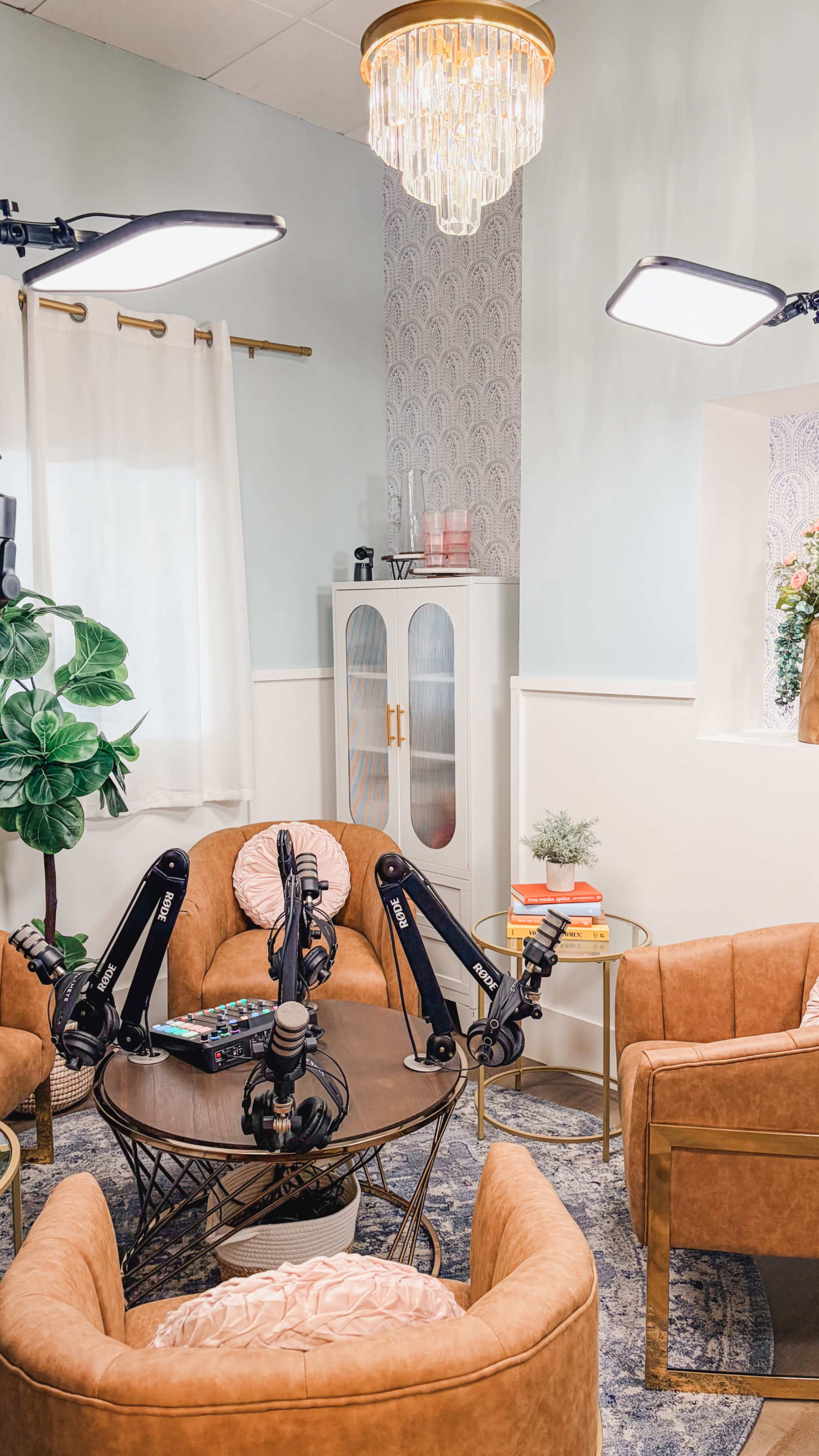 The image shows a cozy podcasting studio with brown chairs arranged around a circular table, surrounded by green plants and illuminated by a chandelier.