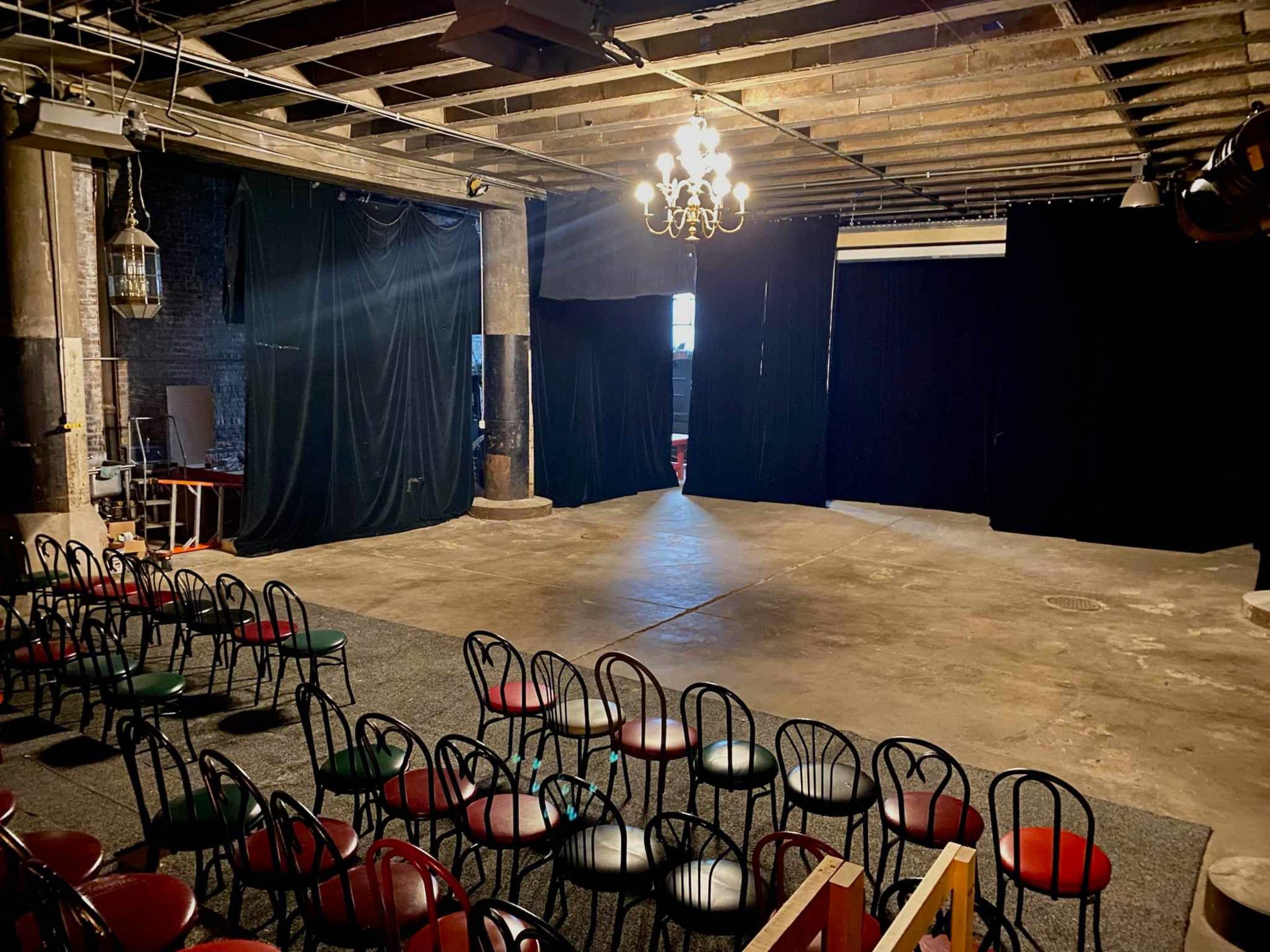 A dimly lit performance space features empty rows of chairs facing a curtained stage area.