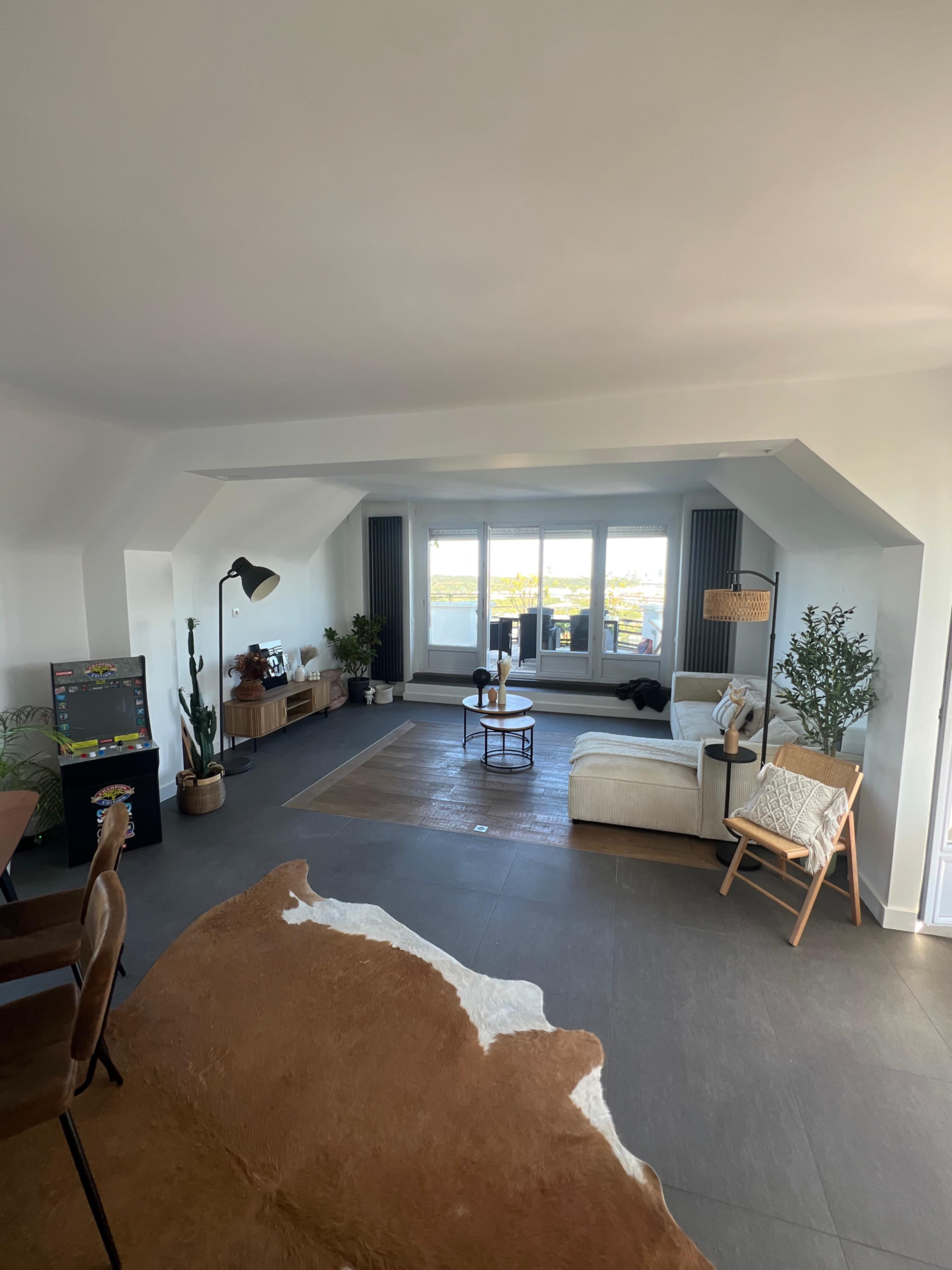 The image shows a spacious living room with large windows, a sectional sofa, a coffee table, and decorative plants.