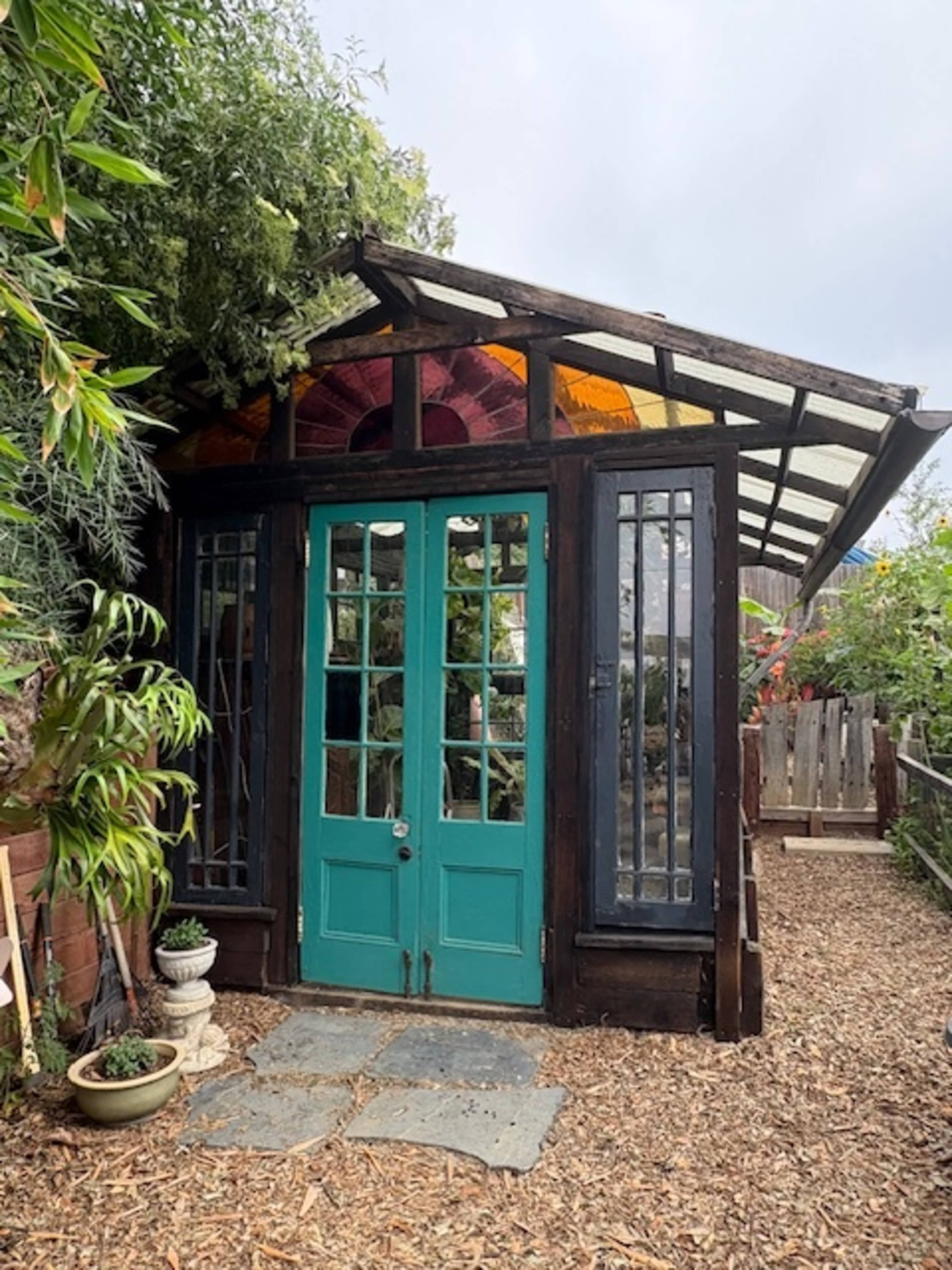 The image shows a small garden shed with teal double doors, large windows, and a slanted glass roof surrounded by greenery.