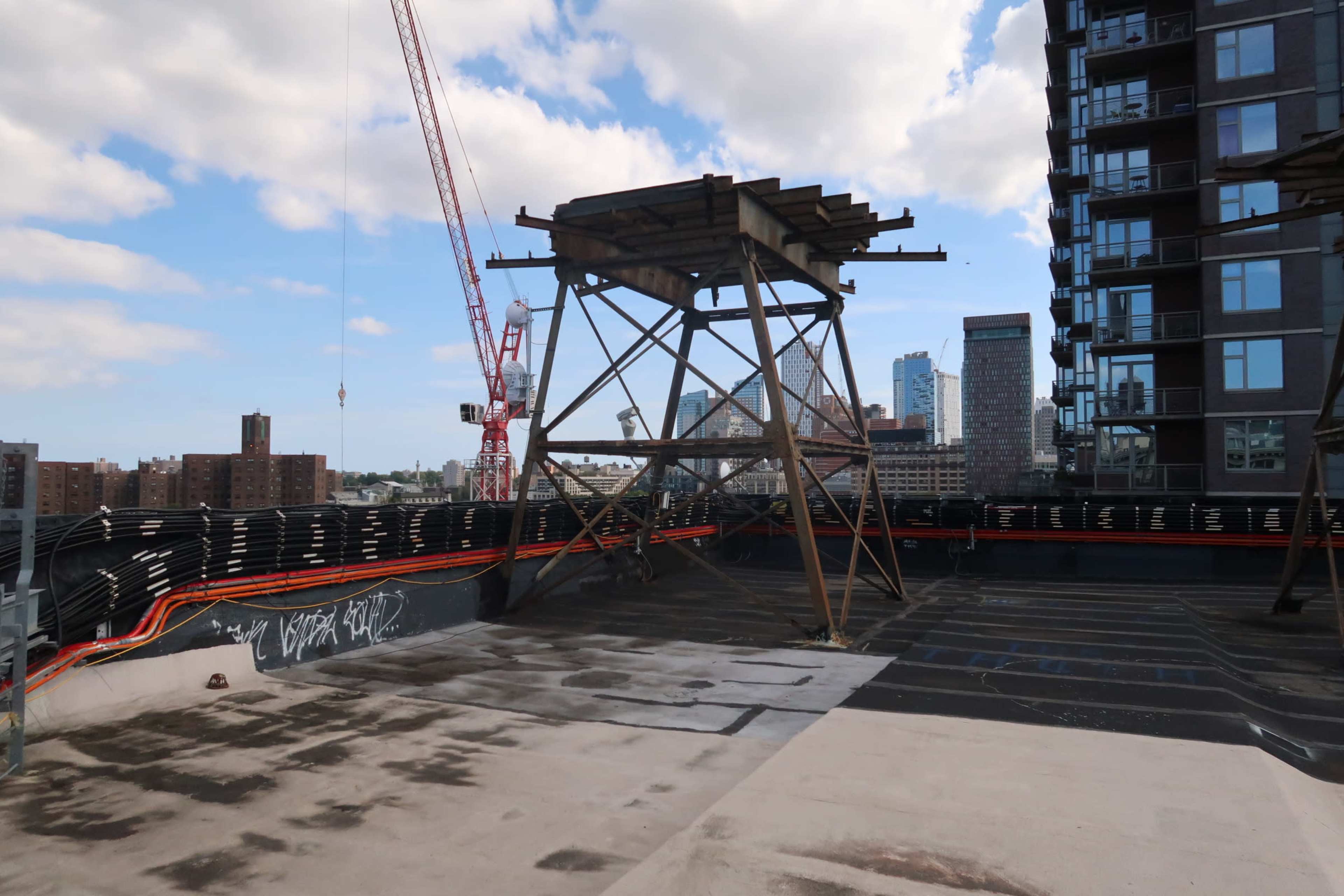 A construction site rooftop featuring a metal framework and a nearby crane, with a view of city buildings in the background.
