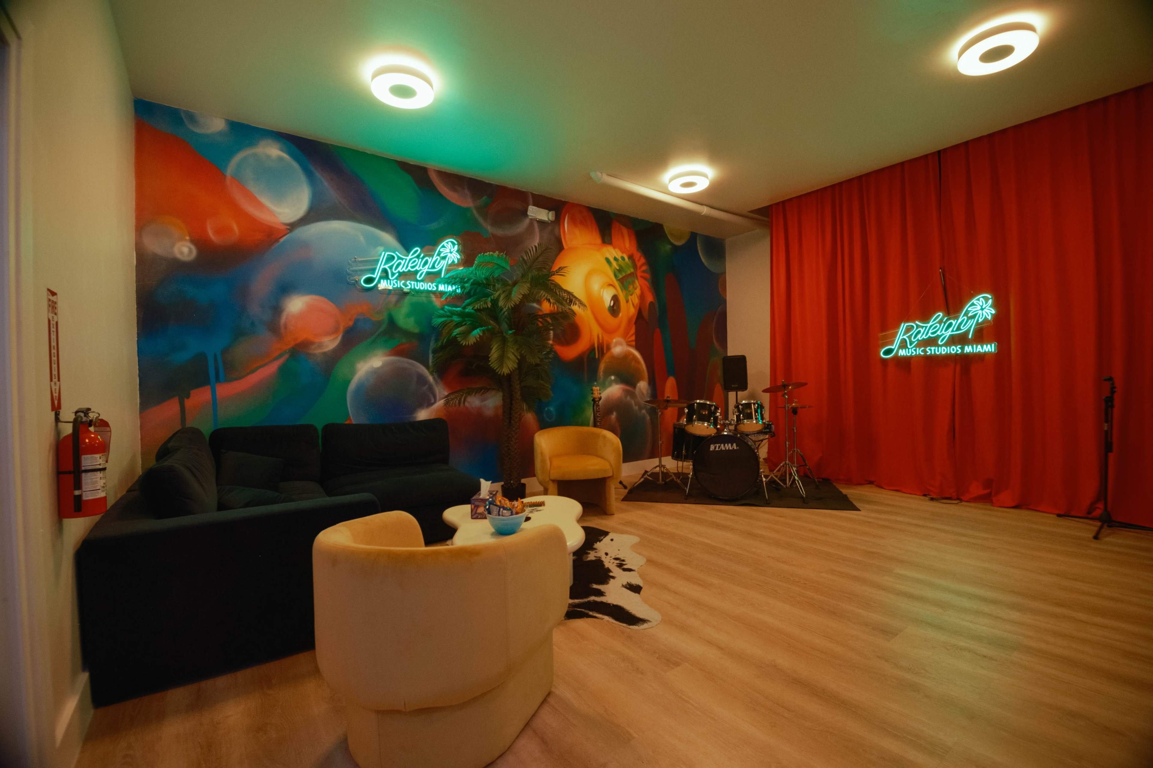 The image shows a music-themed lounge area with a colorful mural, a black sofa, a light yellow armchair, a small table, and a drum set near a red curtain.