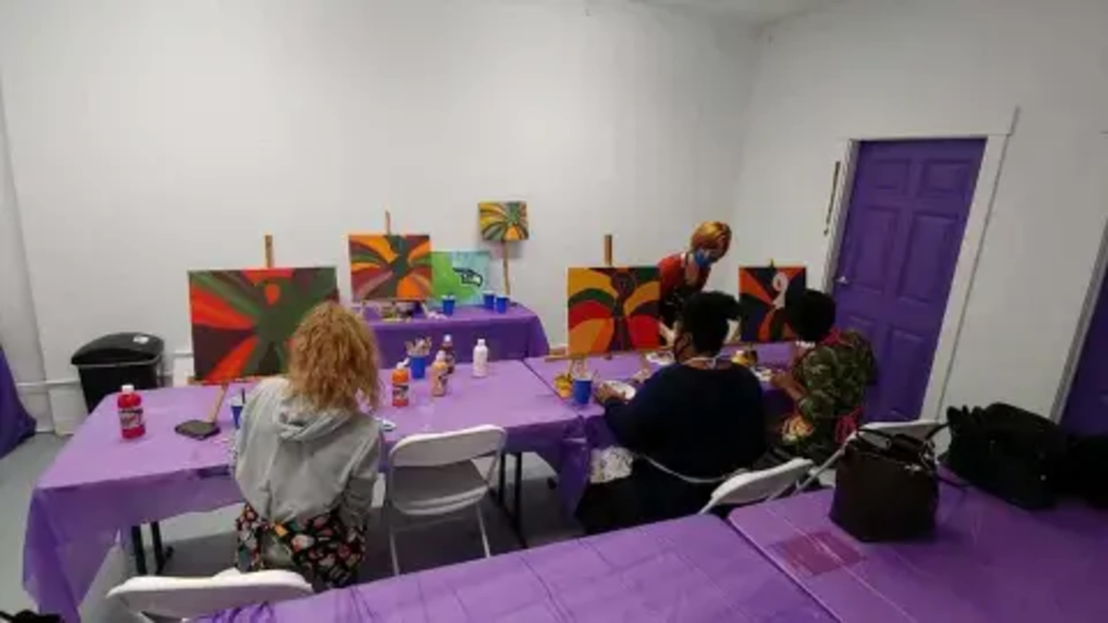 A group of people are painting on canvases at tables covered with purple cloth in a brightly lit room.