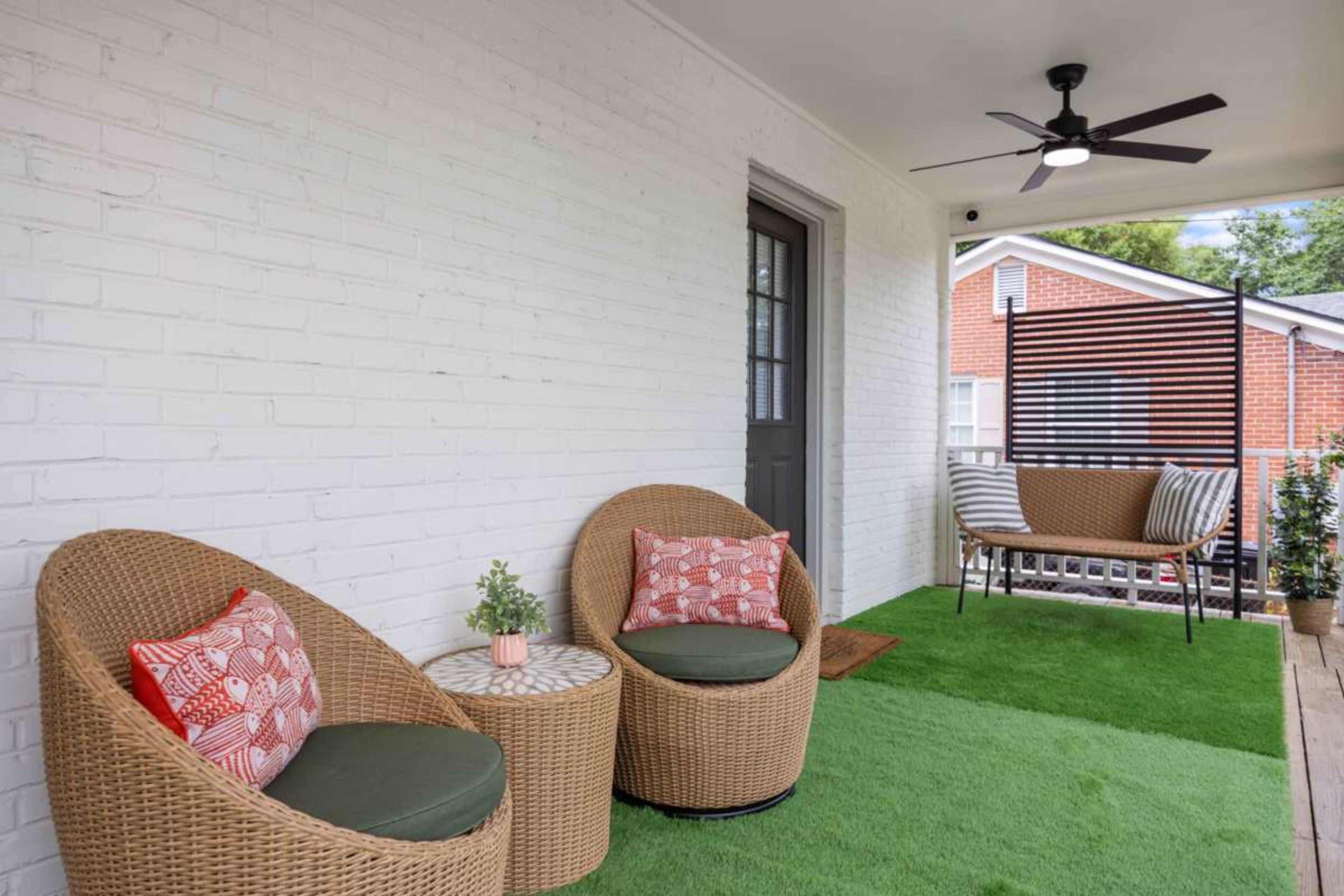 The image shows a cozy porch area featuring two wicker chairs, a side table, and a bench, with green carpet and a white brick wall.