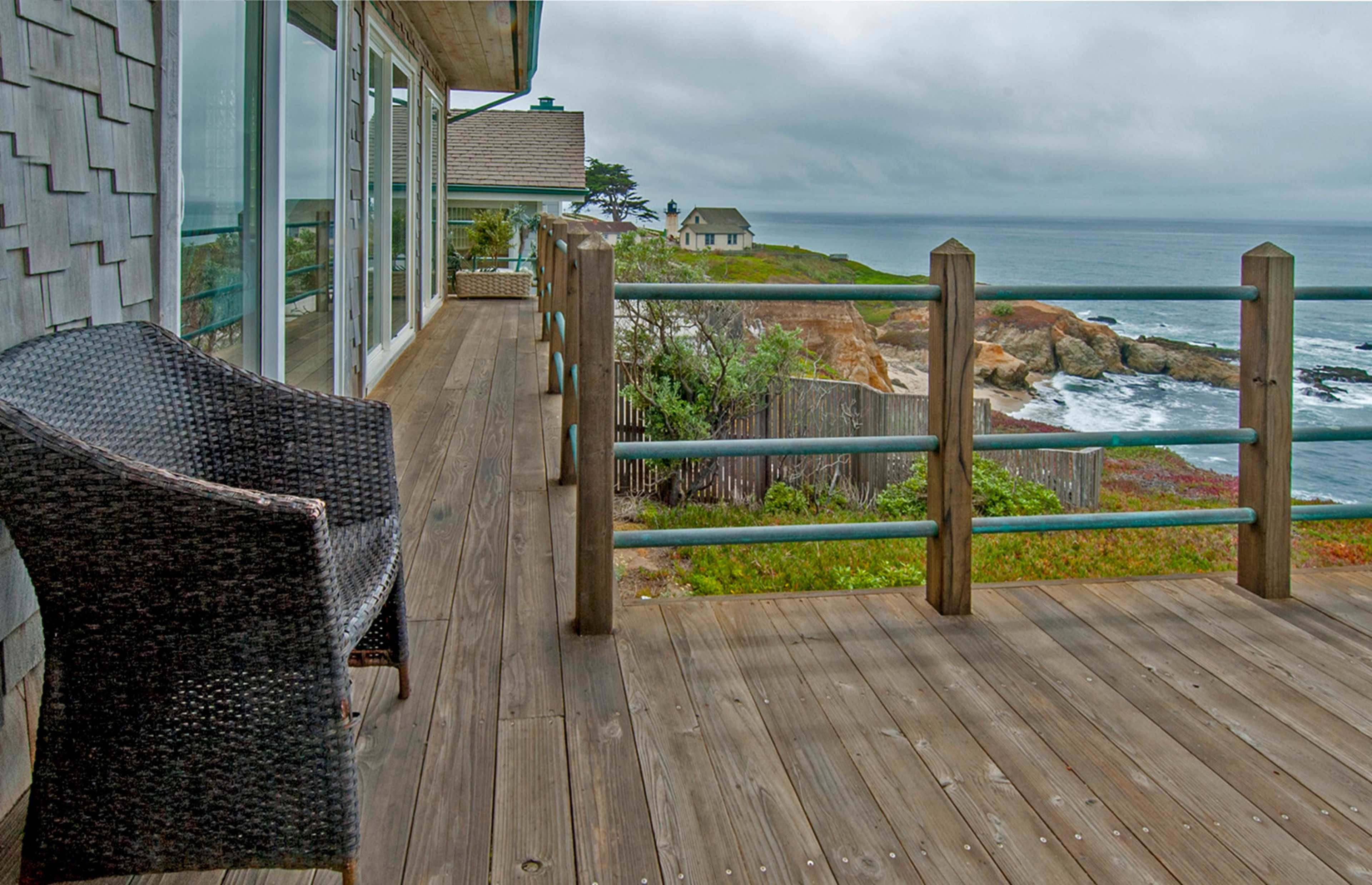 A wooden deck with a wicker chair overlooks a rocky coastline under a cloudy sky.