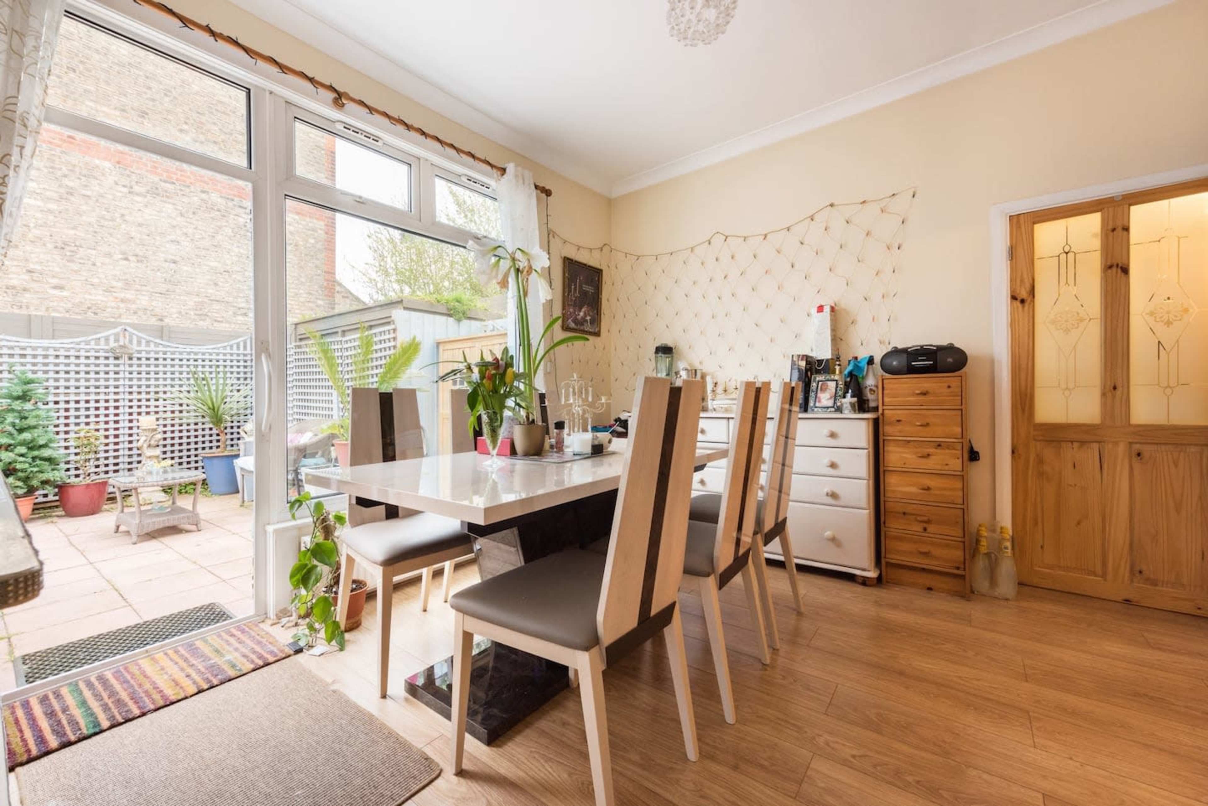 A bright dining area features a large table surrounded by upright chairs, with a view of an outdoor patio and greenery through large windows.
