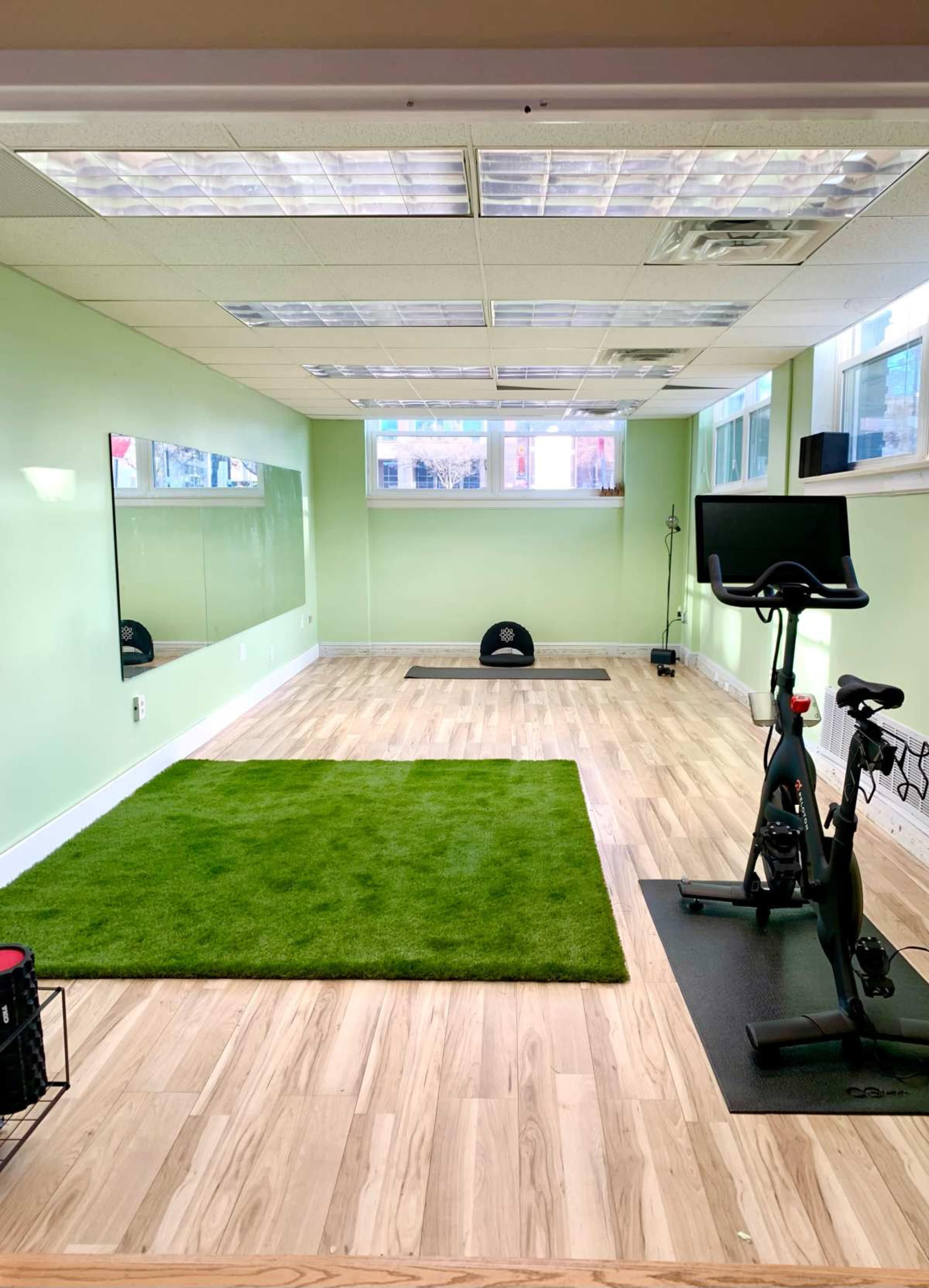 The image shows a bright, empty workout room with green walls, large windows, a mirrored wall, a stationary bike, and a green exercise mat on the wooden floor.