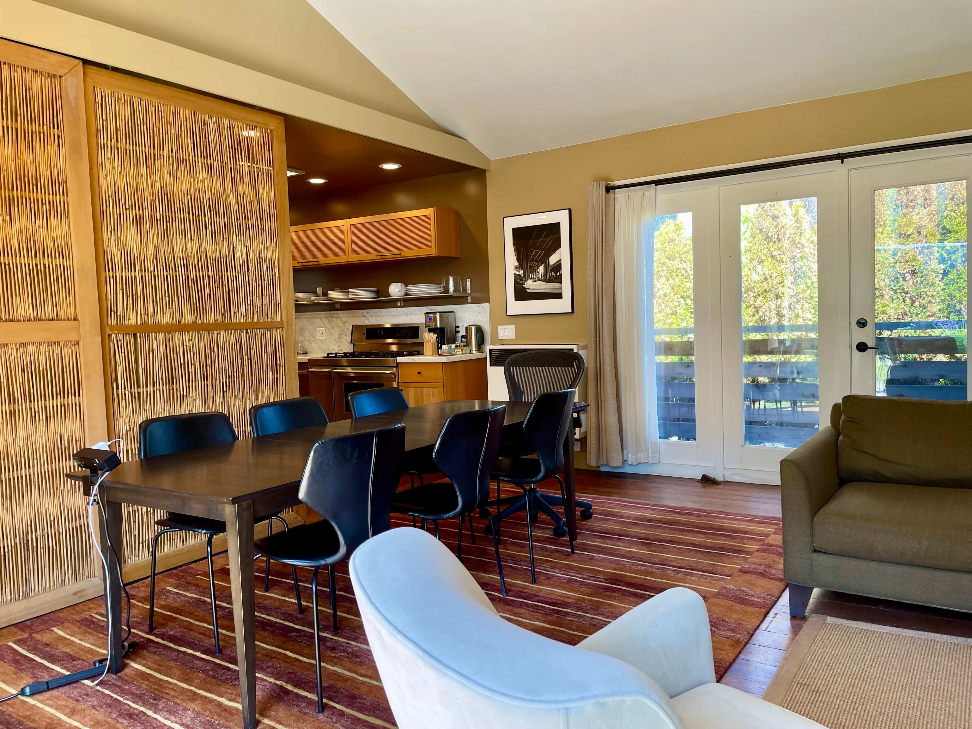 The image shows a modern dining area with a wooden table and black chairs, adjacent to a kitchen and featuring sliding glass doors that open to a balcony.