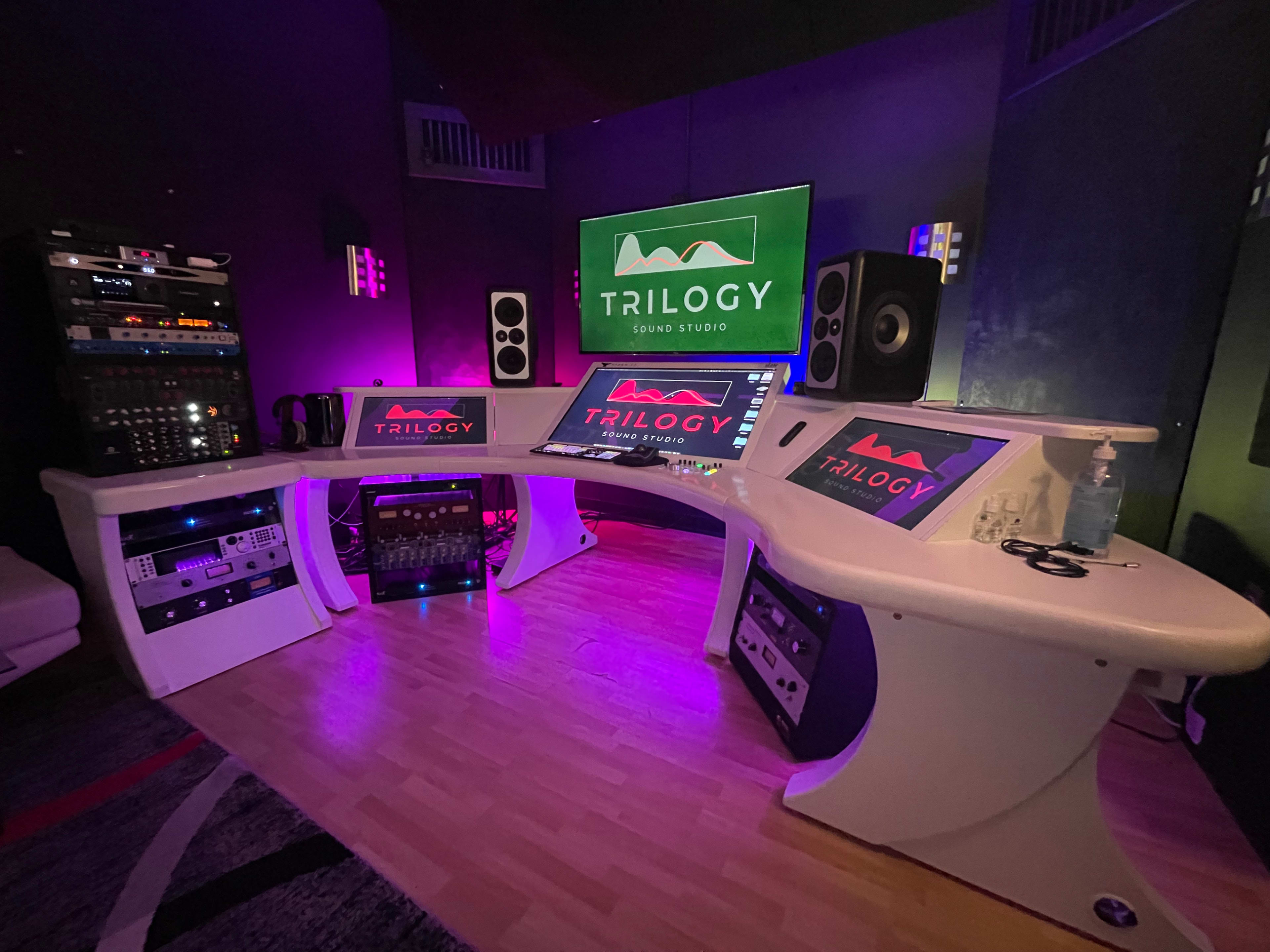 The image shows a modern sound studio with three curved workstations, multiple audio equipment racks, and colorful lighting.