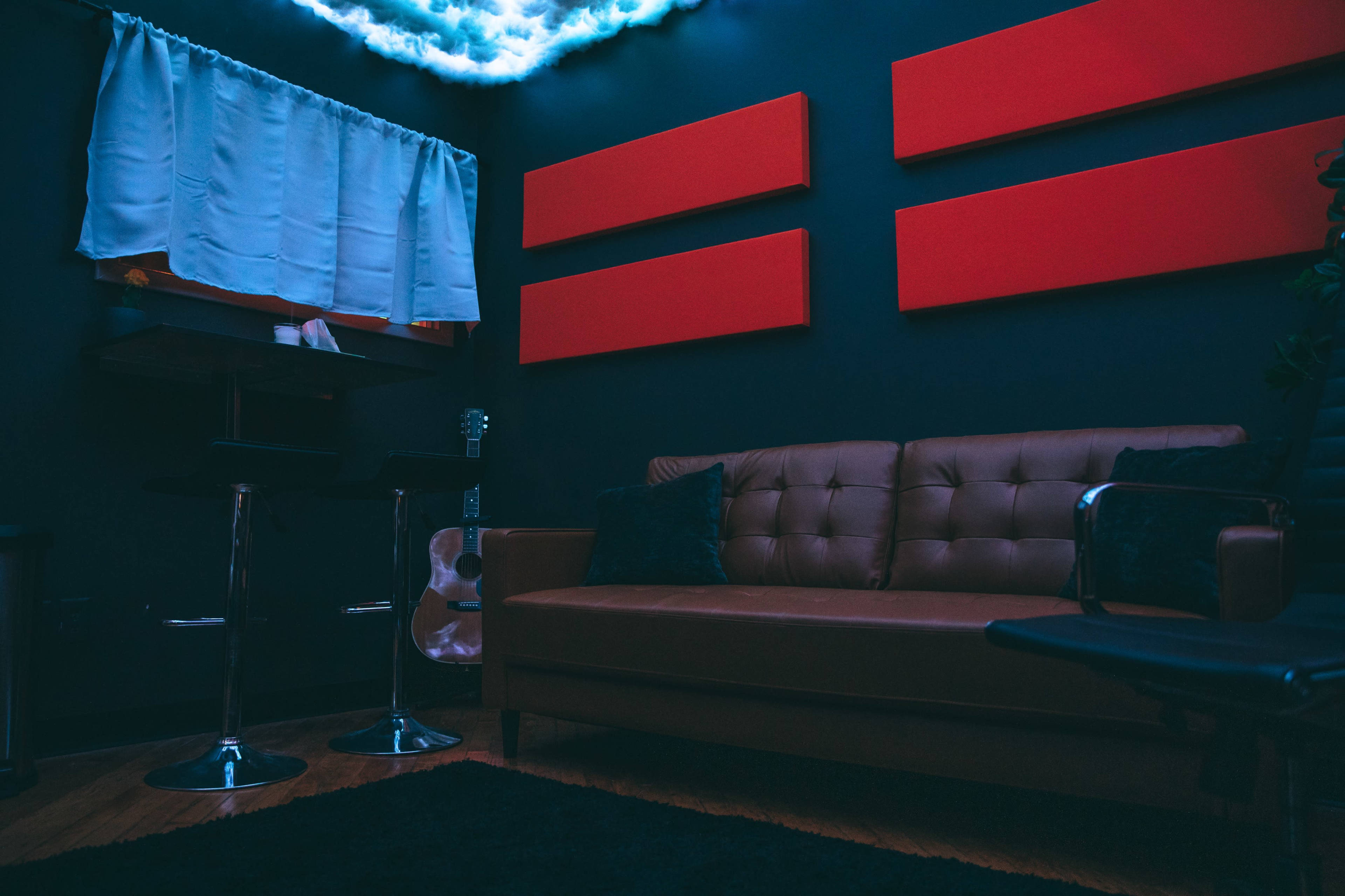 A cozy room with a brown leather sofa, two tall stools, and red acoustic panels on the walls, illuminated by soft lighting.