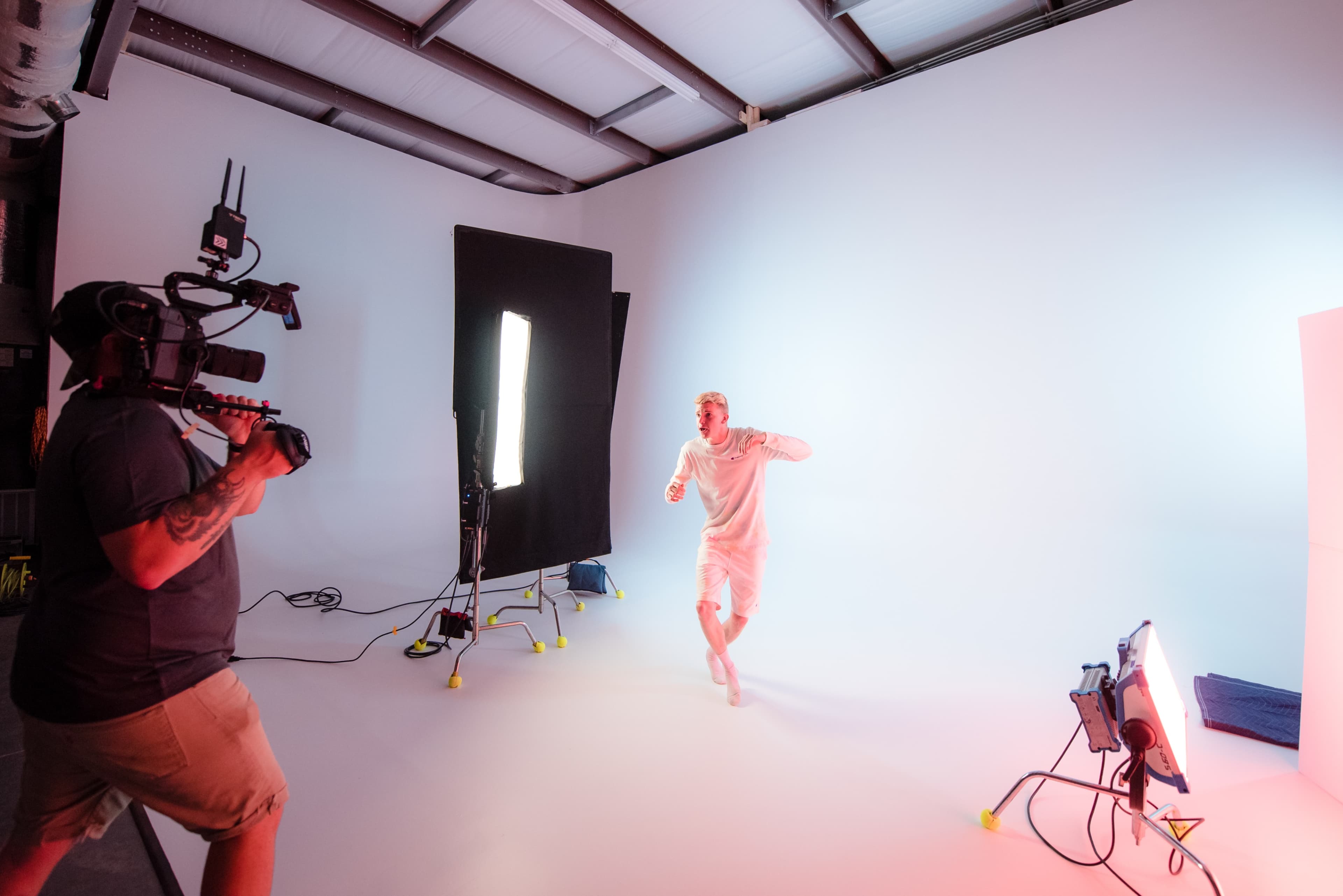 A person in a light-colored outfit is running in a brightly lit photography studio while being filmed by another person holding a camera.