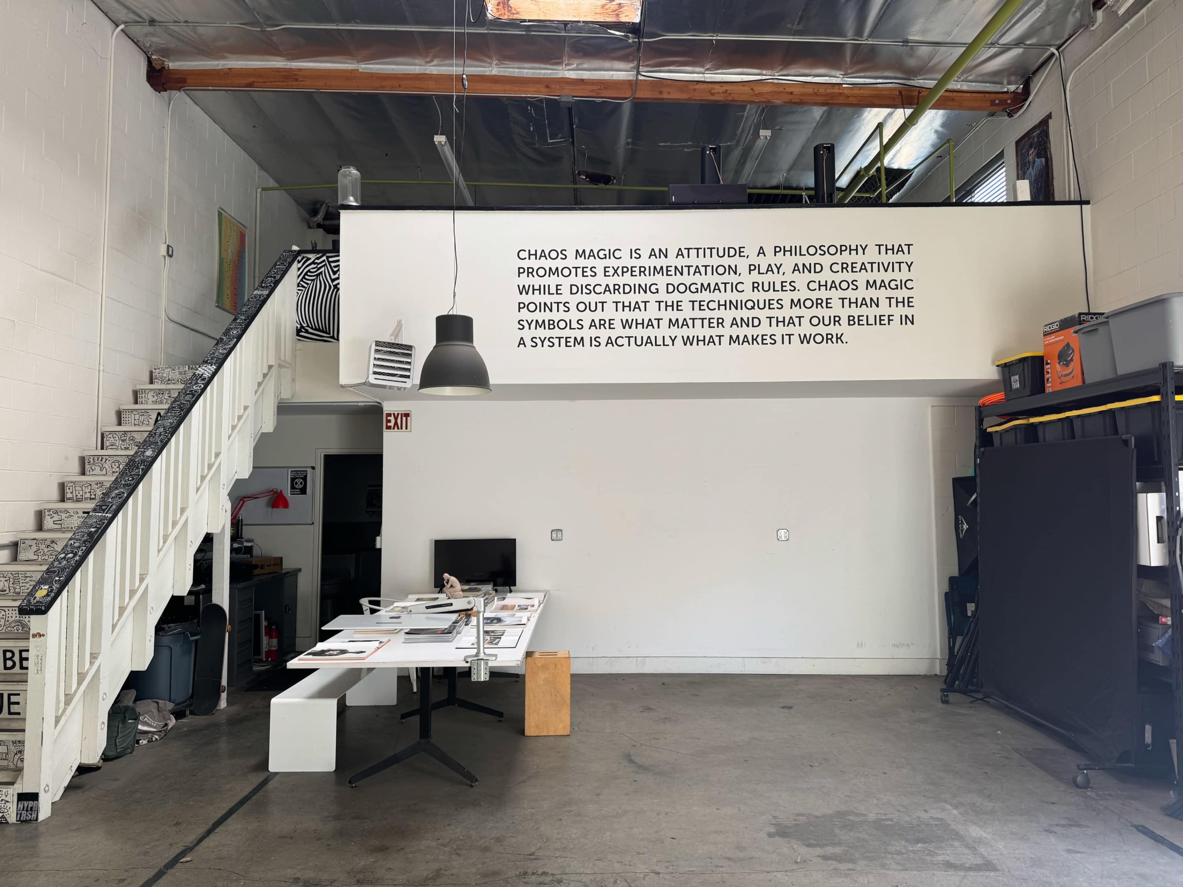 The image shows an industrial interior space with a staircase leading to a loft, where a large text about chaos magic is displayed on the wall above a desk filled with papers and materials.