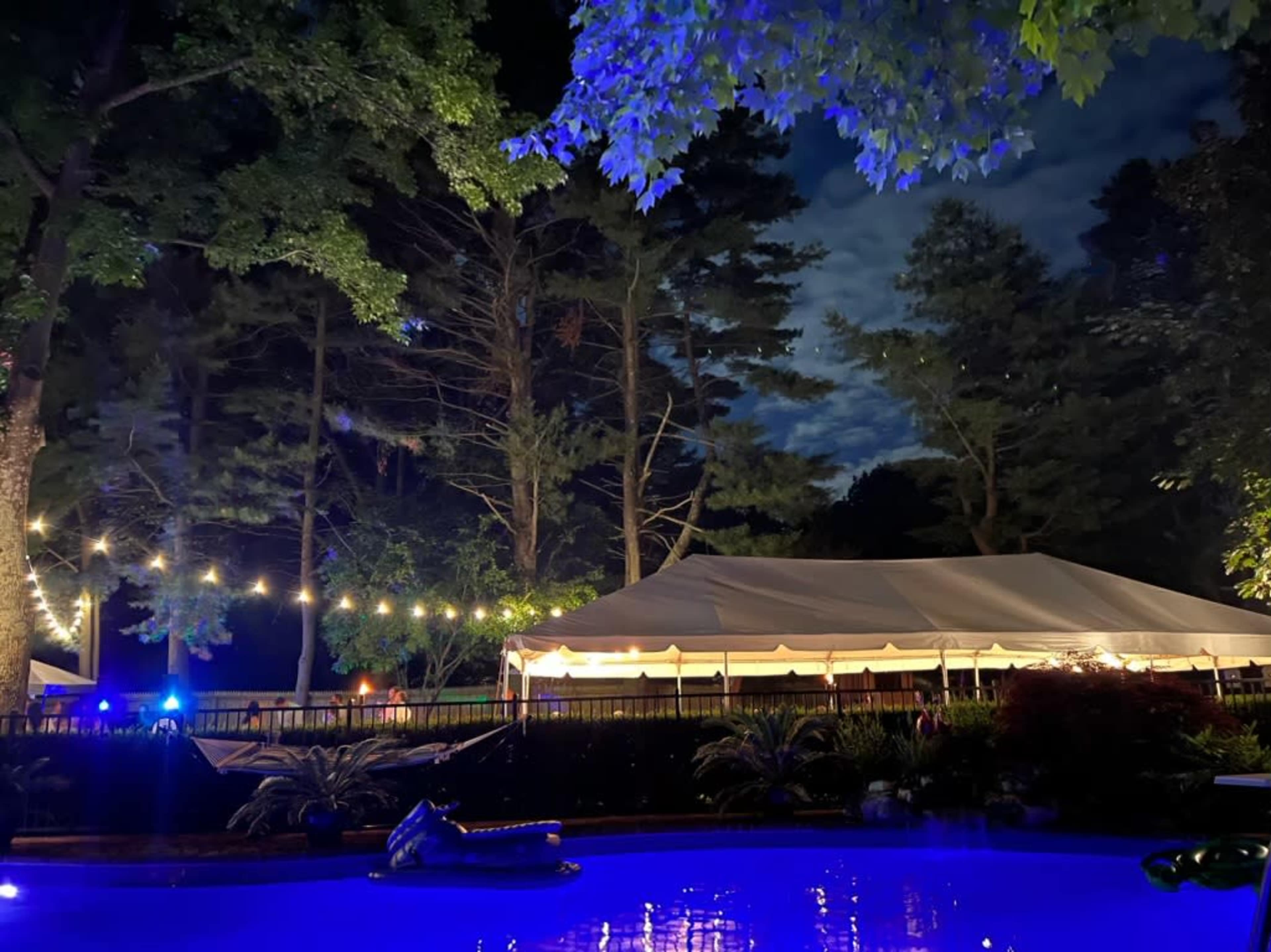 A large white tent is set up beside a pool, illuminated by string lights against a backdrop of dark trees and a night sky.