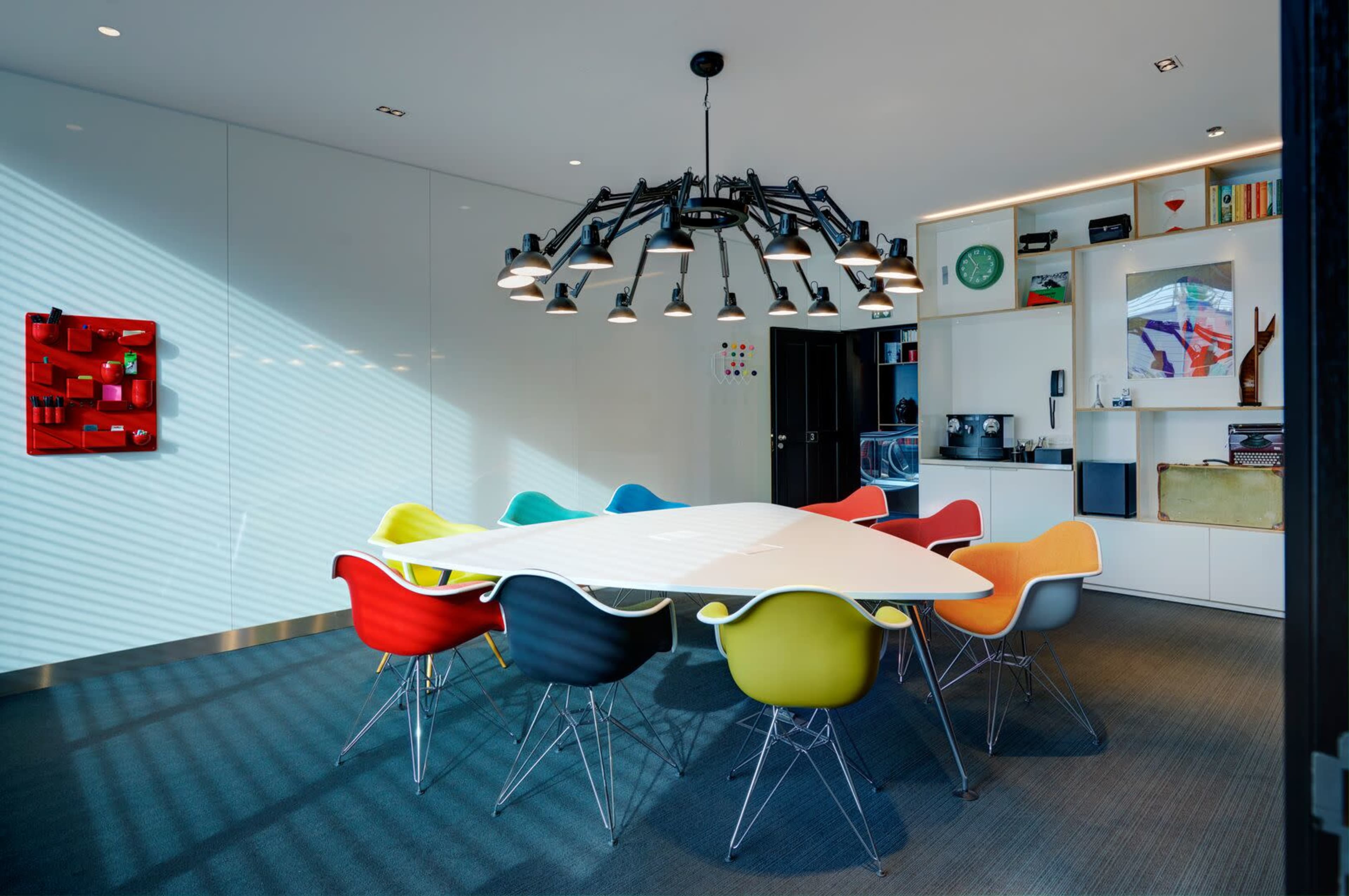 A modern conference room features a large triangular table surrounded by colorful chairs and a striking chandelier above.