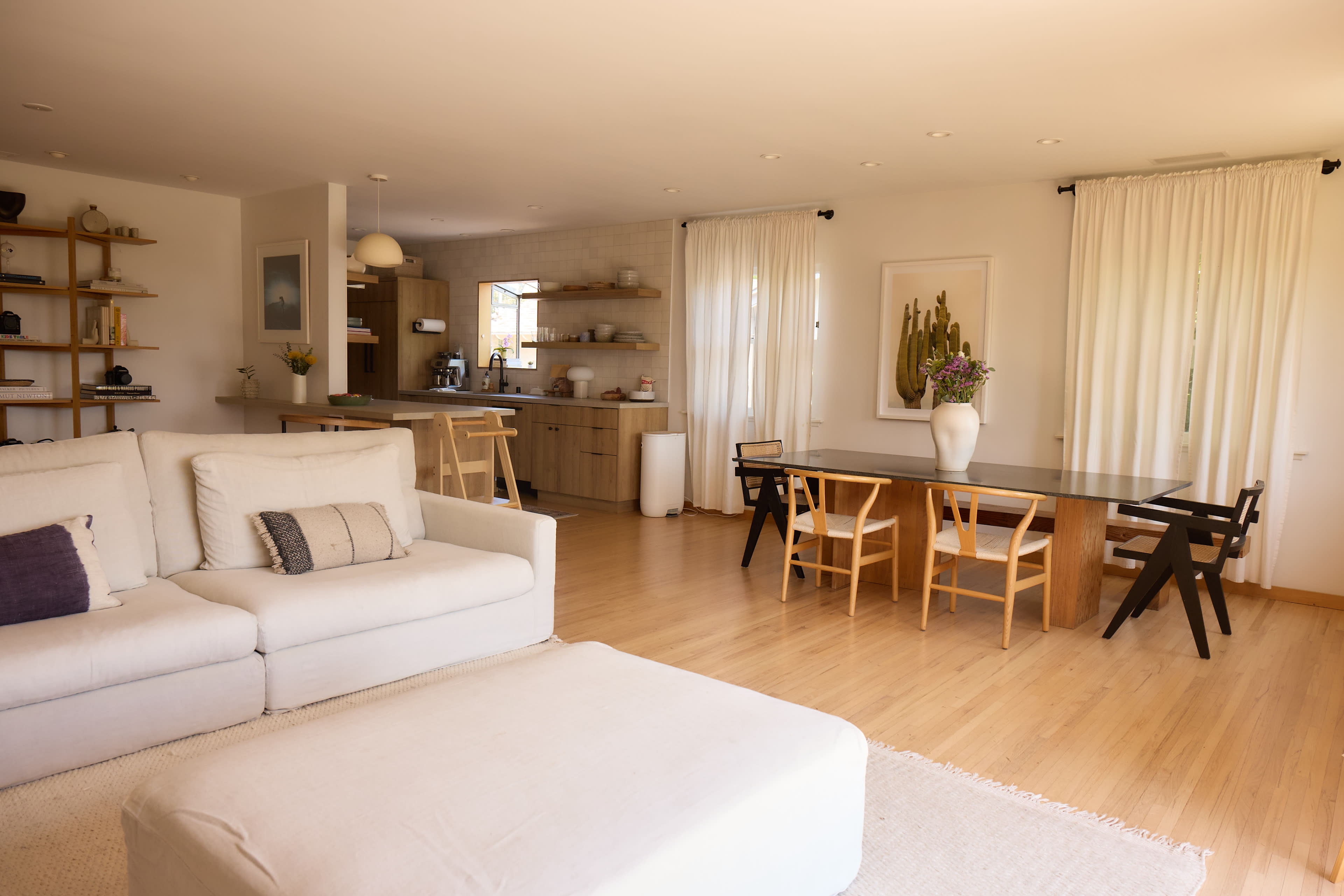 A spacious living area features a white sofa, a dining table with wooden chairs, and large windows with sheer curtains.