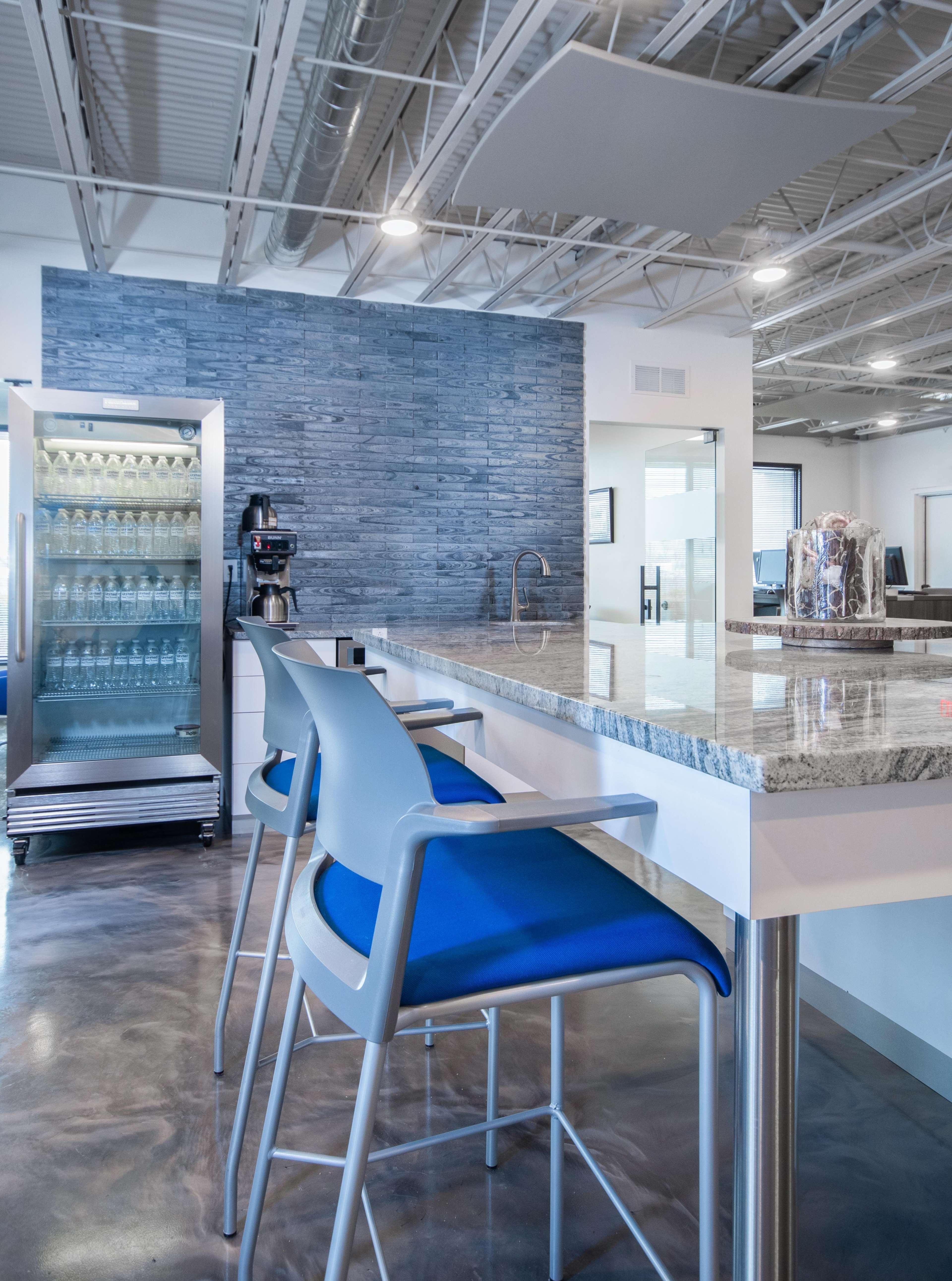 A modern office break area featuring a granite-topped counter with blue chairs, a refrigerator, and a coffee machine against a textured blue wall.