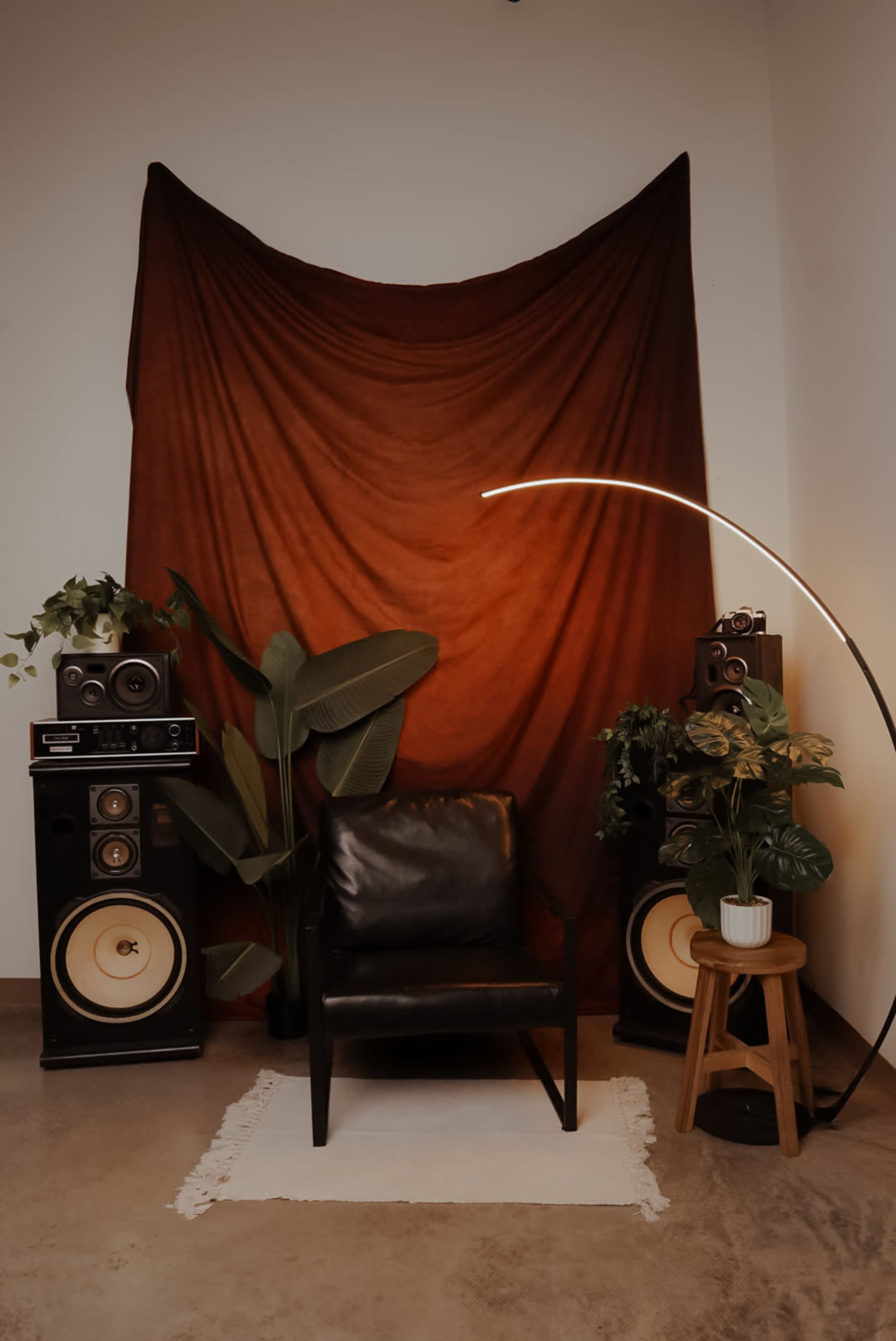 The image shows a cozy interior scene with a black chair in front of a rust-colored backdrop, flanked by two vintage speakers and a floor lamp, along with potted plants.