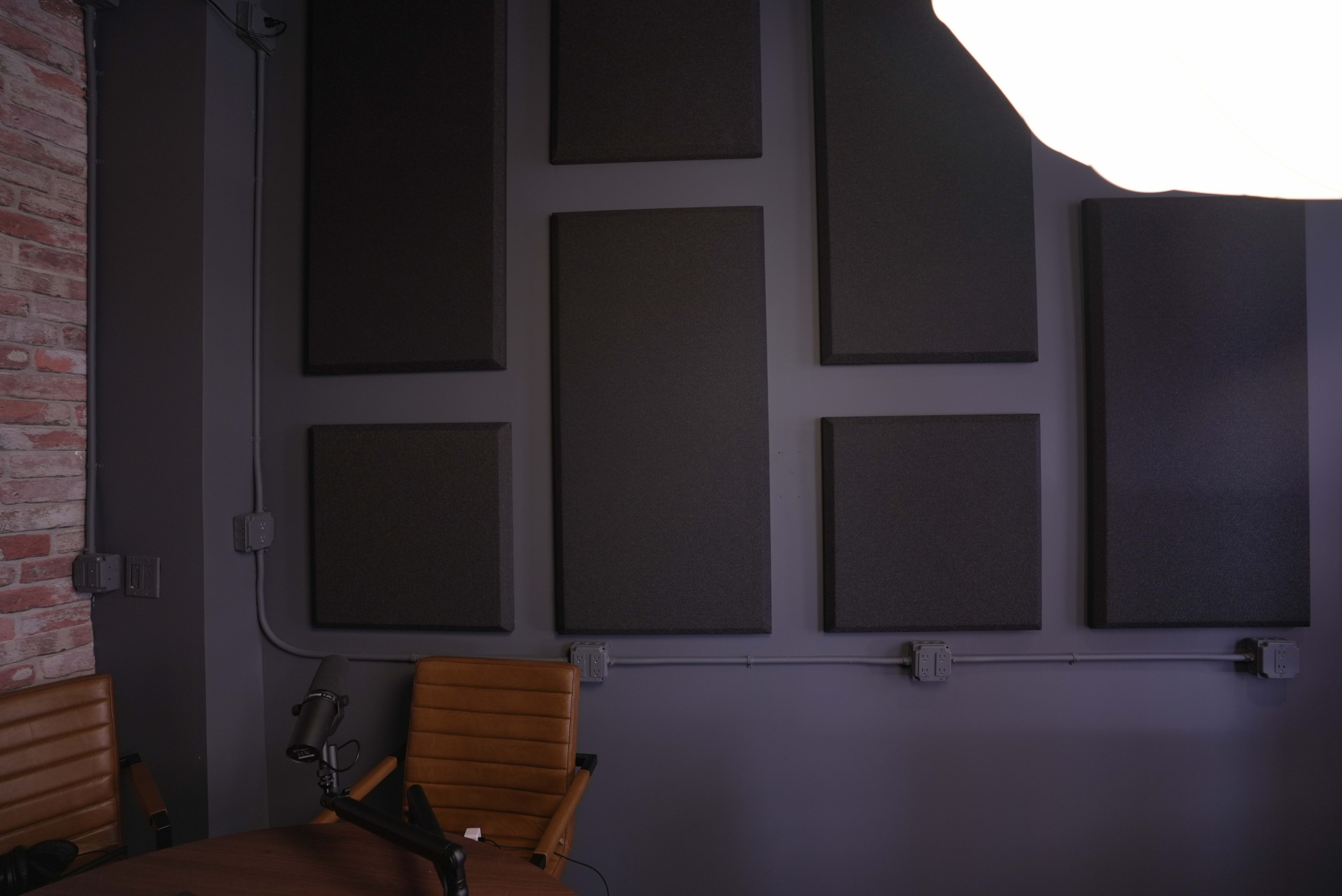 The image shows a wall with several large black acoustic panels mounted on it, next to a round wooden table and two brown leather chairs.