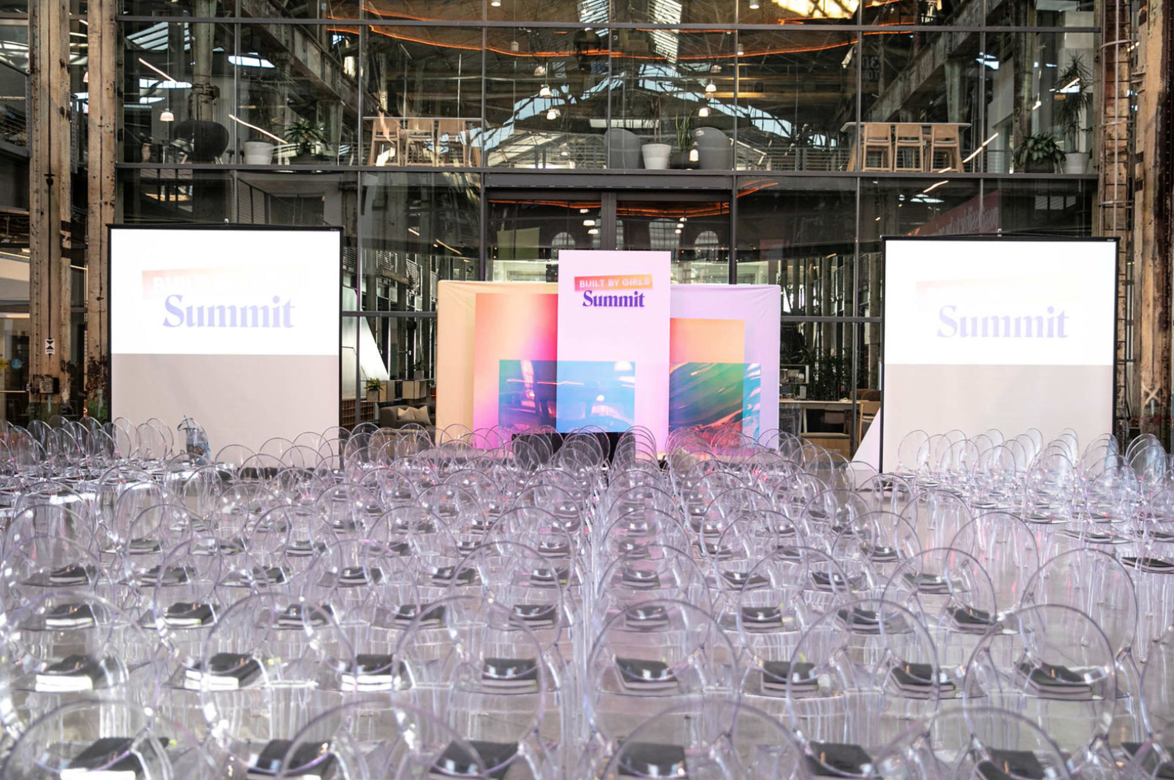 A large hall set up for a summit, with rows of clear chairs facing a stage flanked by two screens displaying the word "Summit."