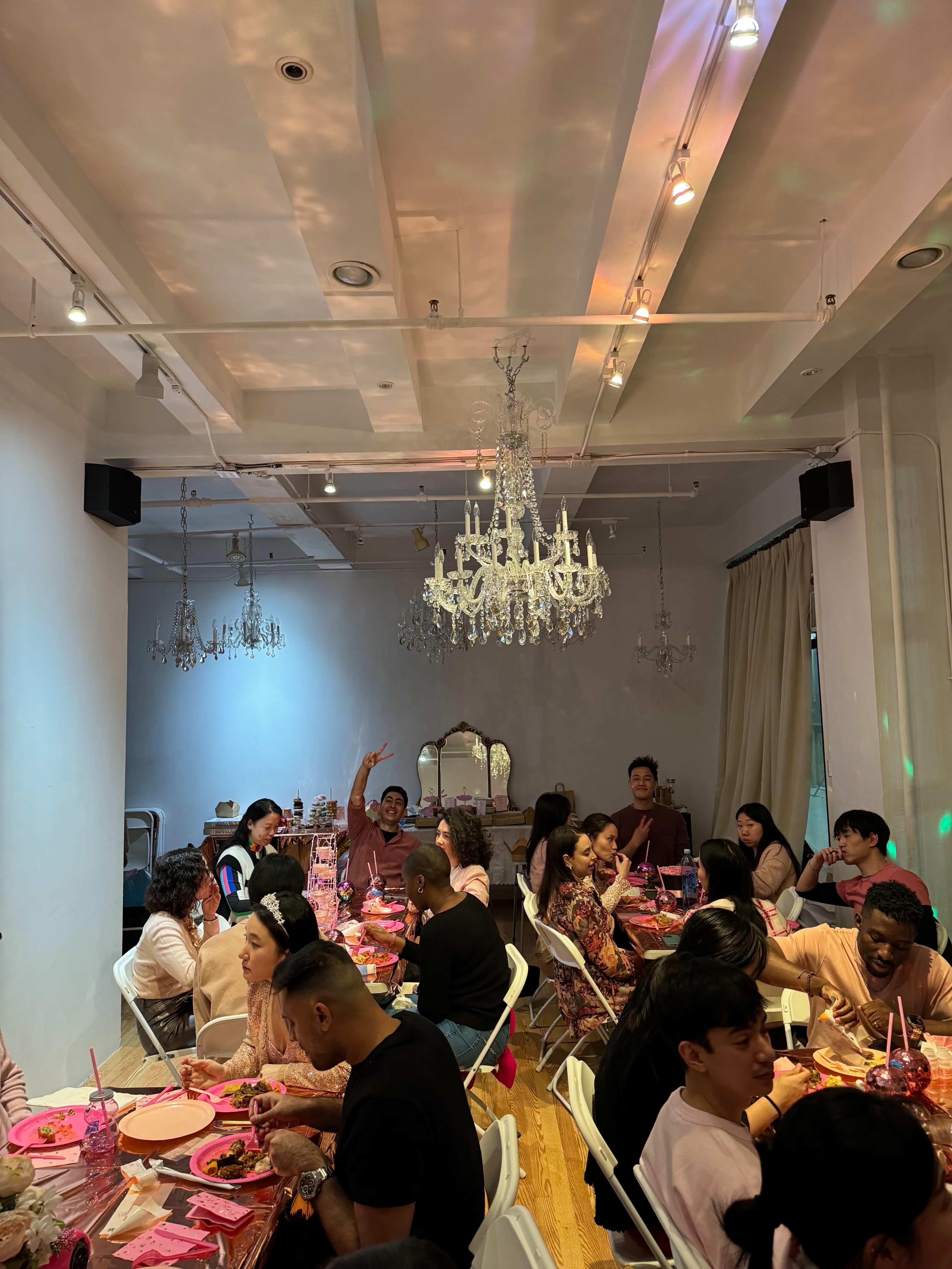 A group of people sits around tables in a brightly lit room, enjoying a meal under a chandelier.