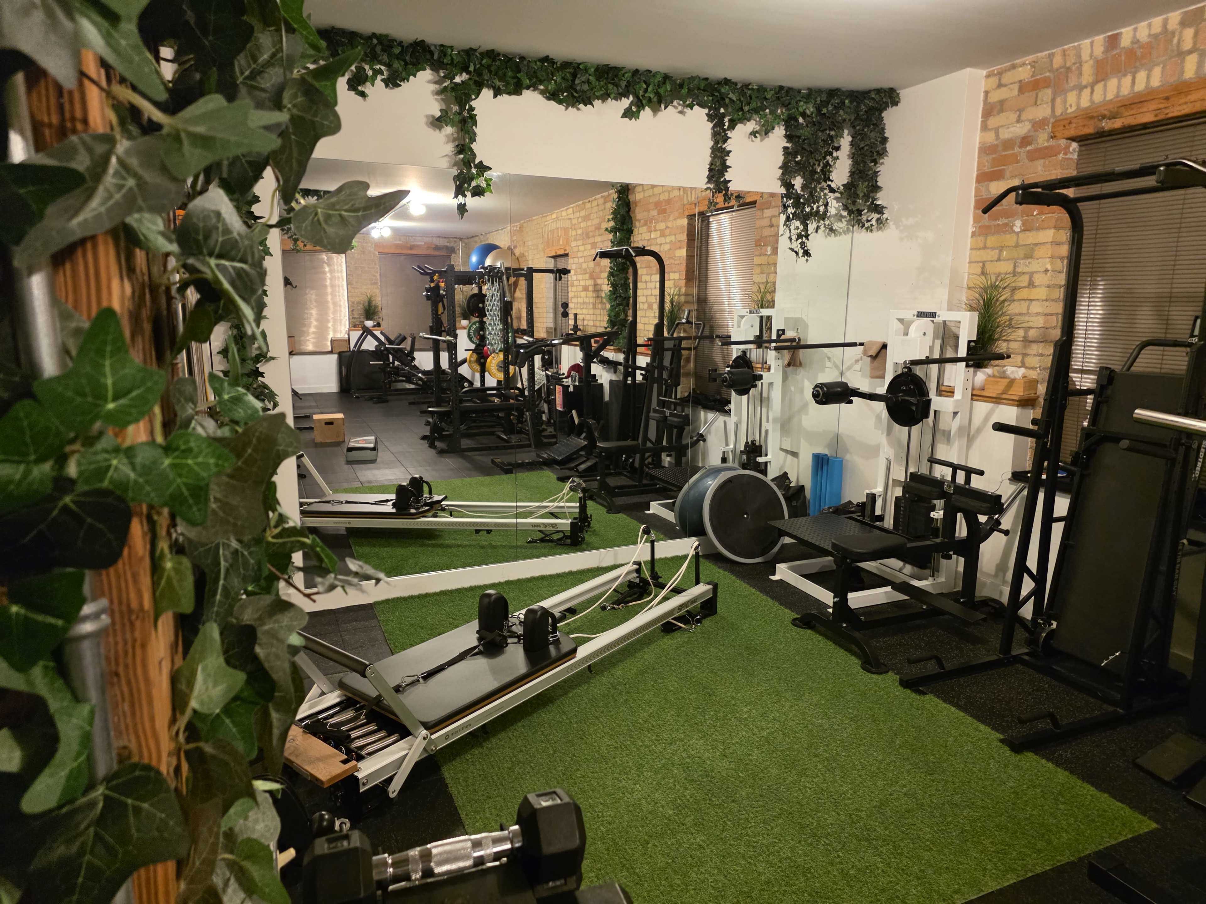 The image shows an indoor gym featuring various workout equipment, including weight machines, benches, and free weights, set on artificial turf and surrounded by greenery.