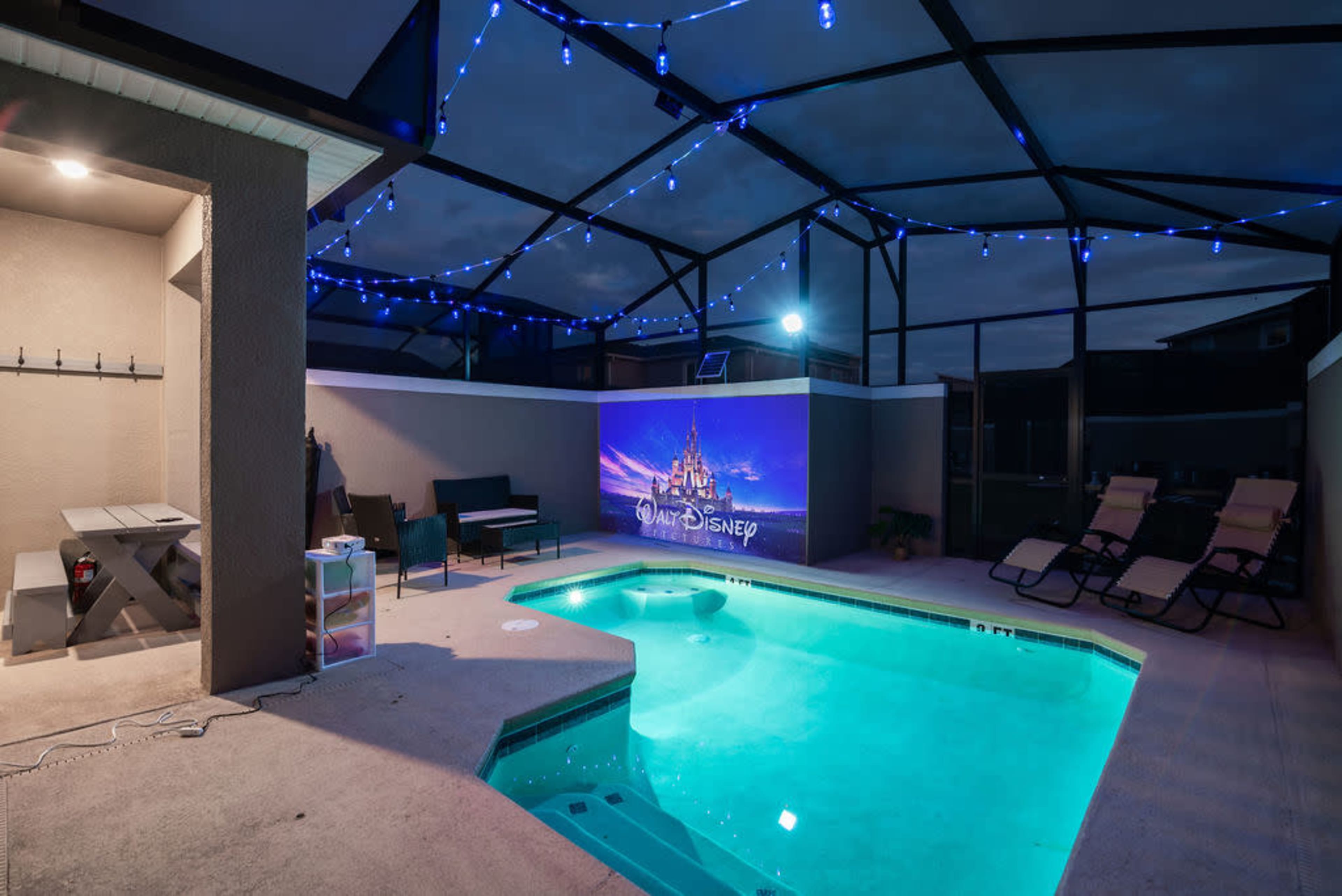 A backyard pool area with a glowing blue pool, a table and chairs, and a large screen displaying a Disney theme set against a nighttime sky.