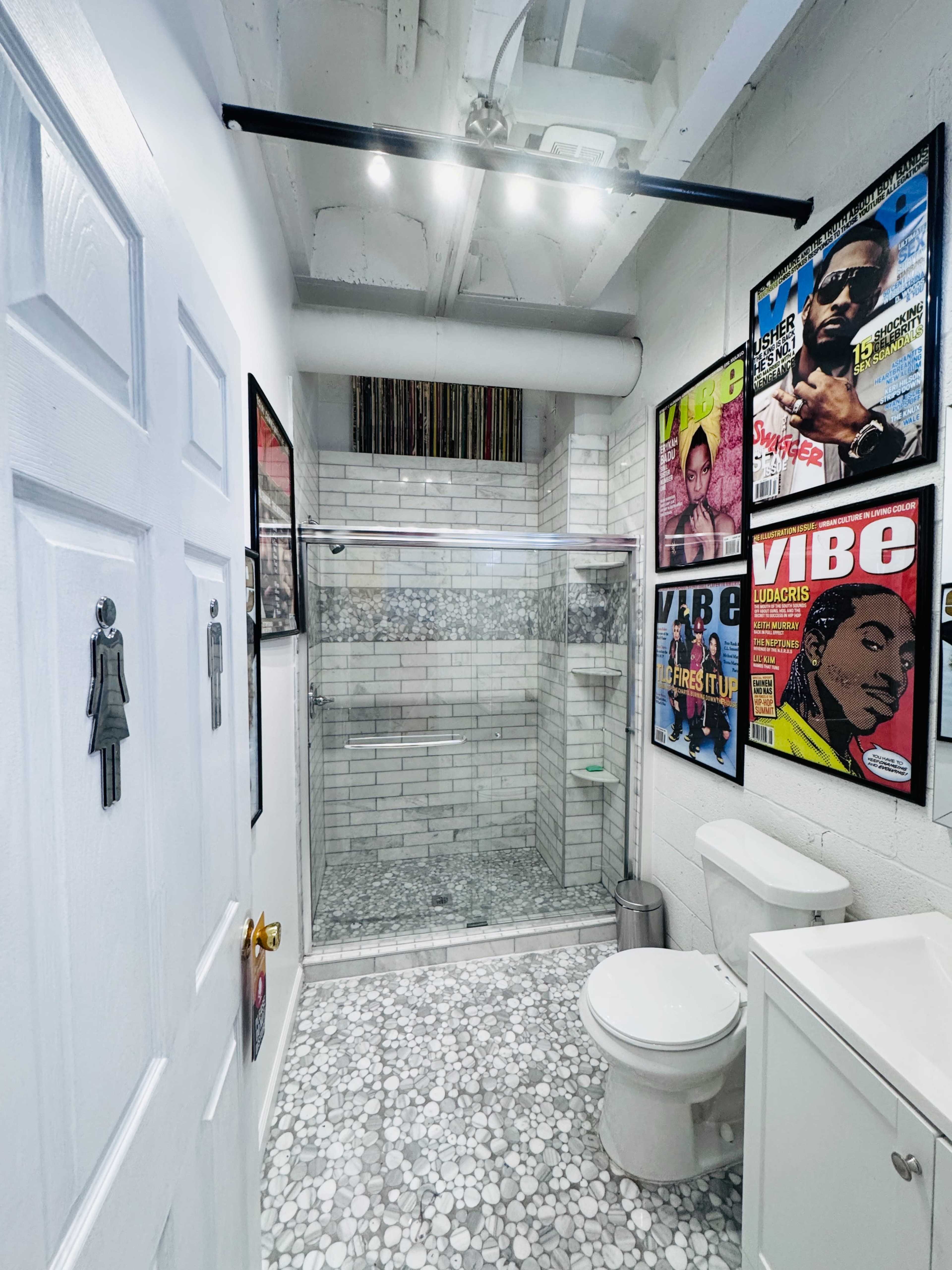 The image shows a modern bathroom featuring white tile walls, a glass shower, a toilet, and a small cabinet, with framed magazine covers on the walls.