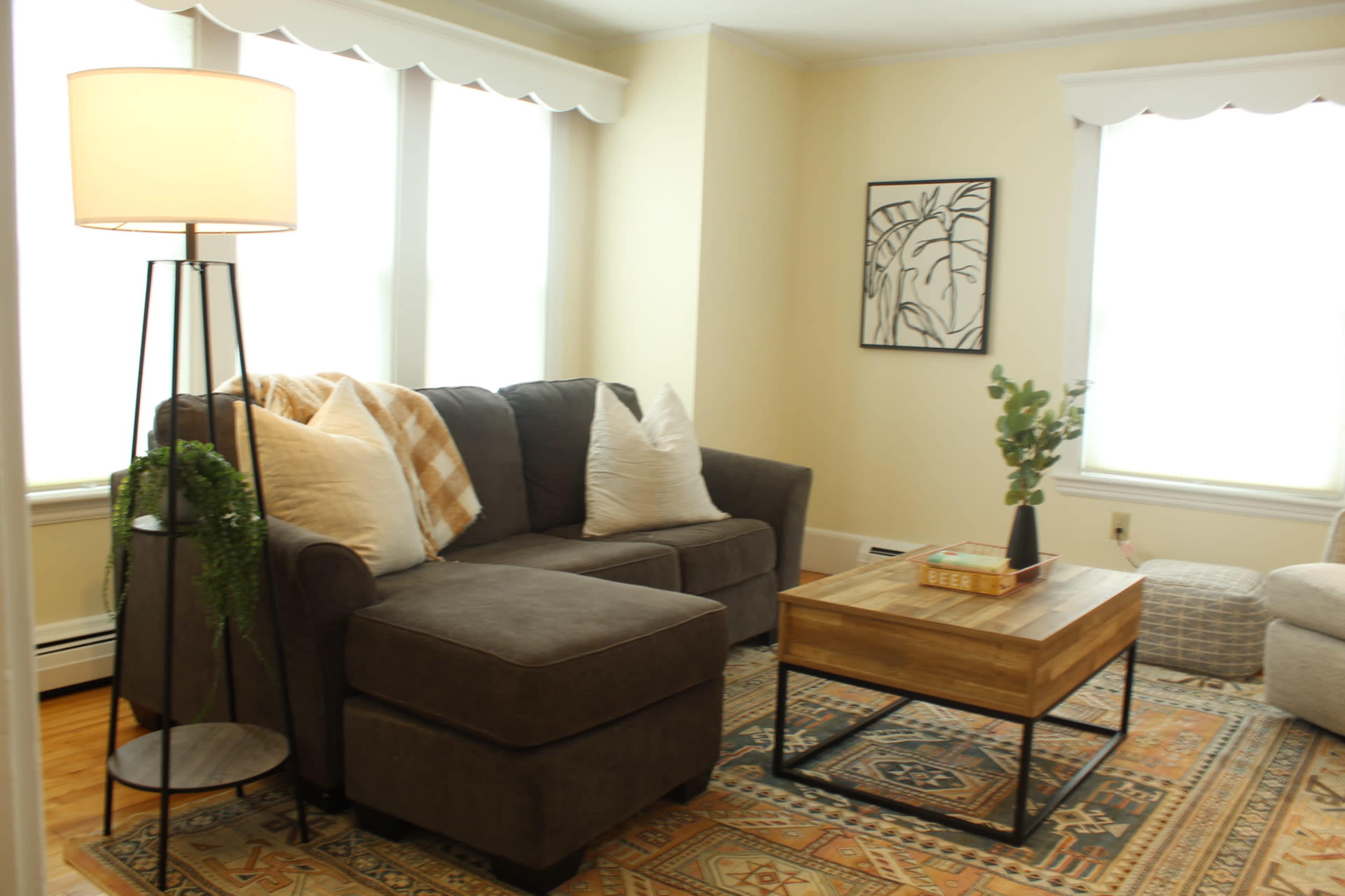 A cozy living room with a gray sectional sofa, a wooden coffee table, and light-colored walls, featuring two large windows and a floor lamp.