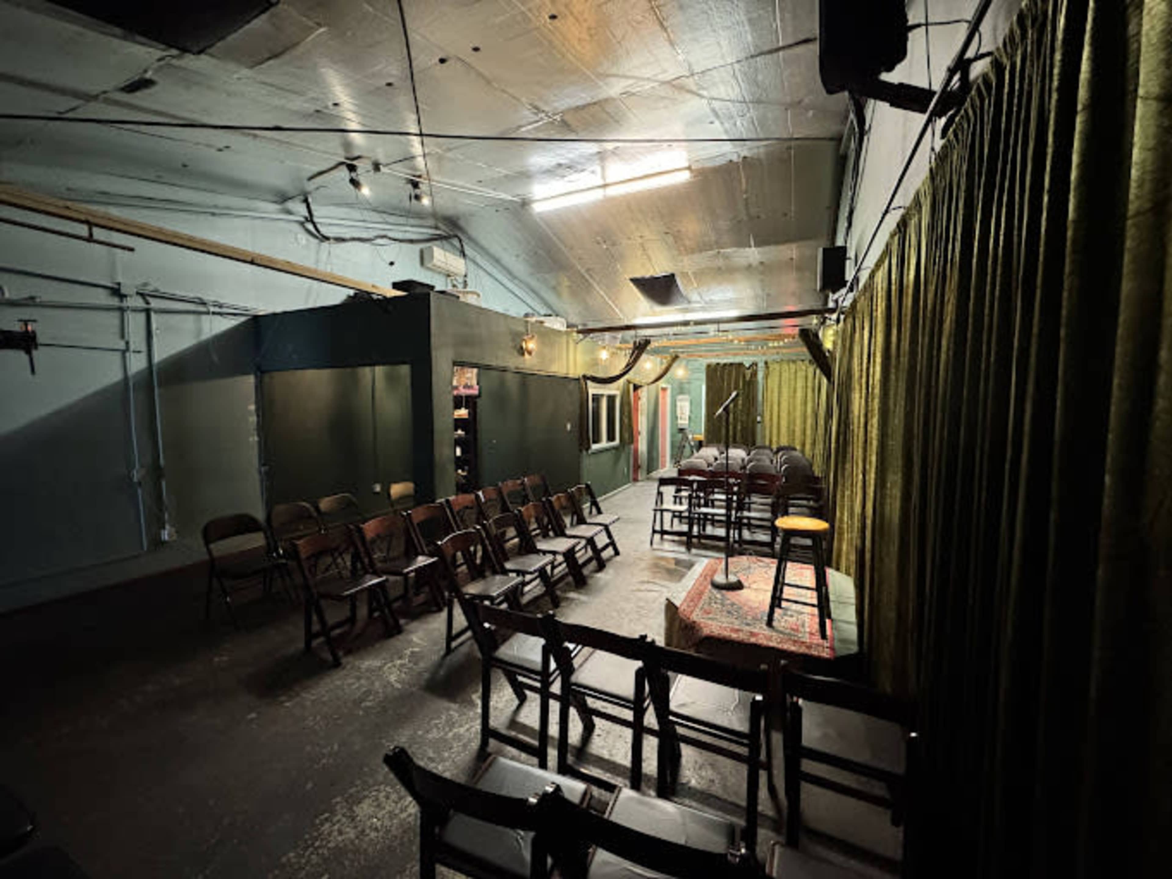 The image shows a dimly lit performance space with rows of folding chairs facing a small stage adorned with a microphone.