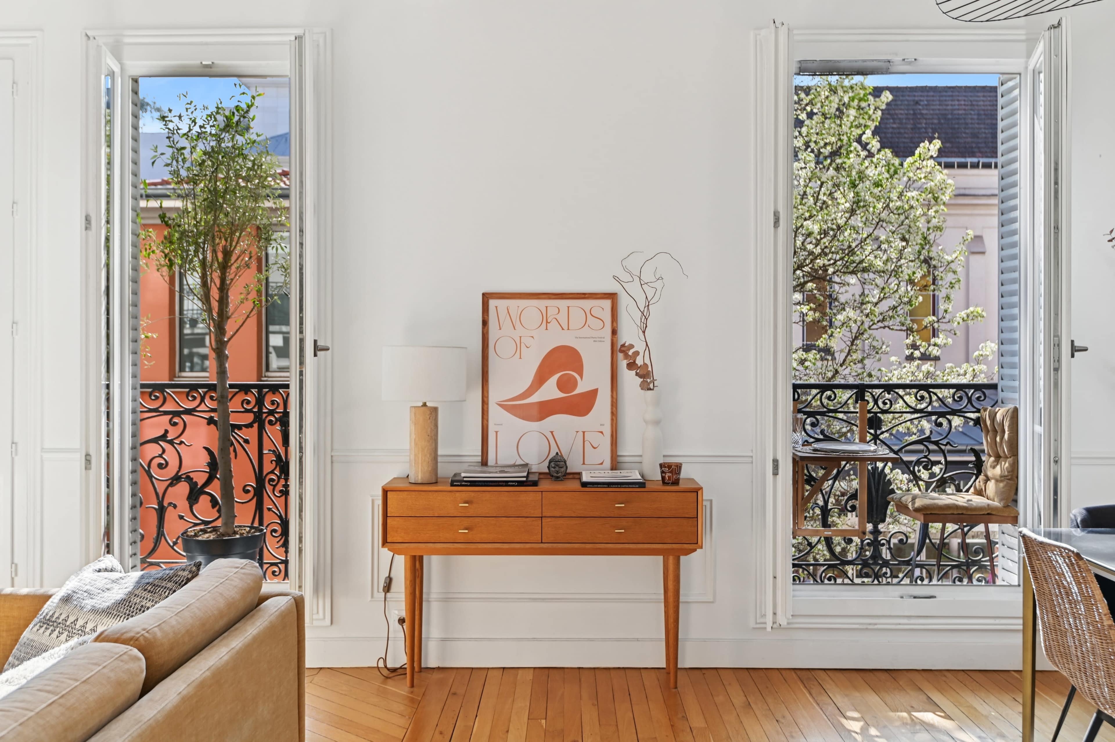 The image shows a room with large windows on either side, featuring a wooden console table adorned with decorative items, and potted plants visible outside.