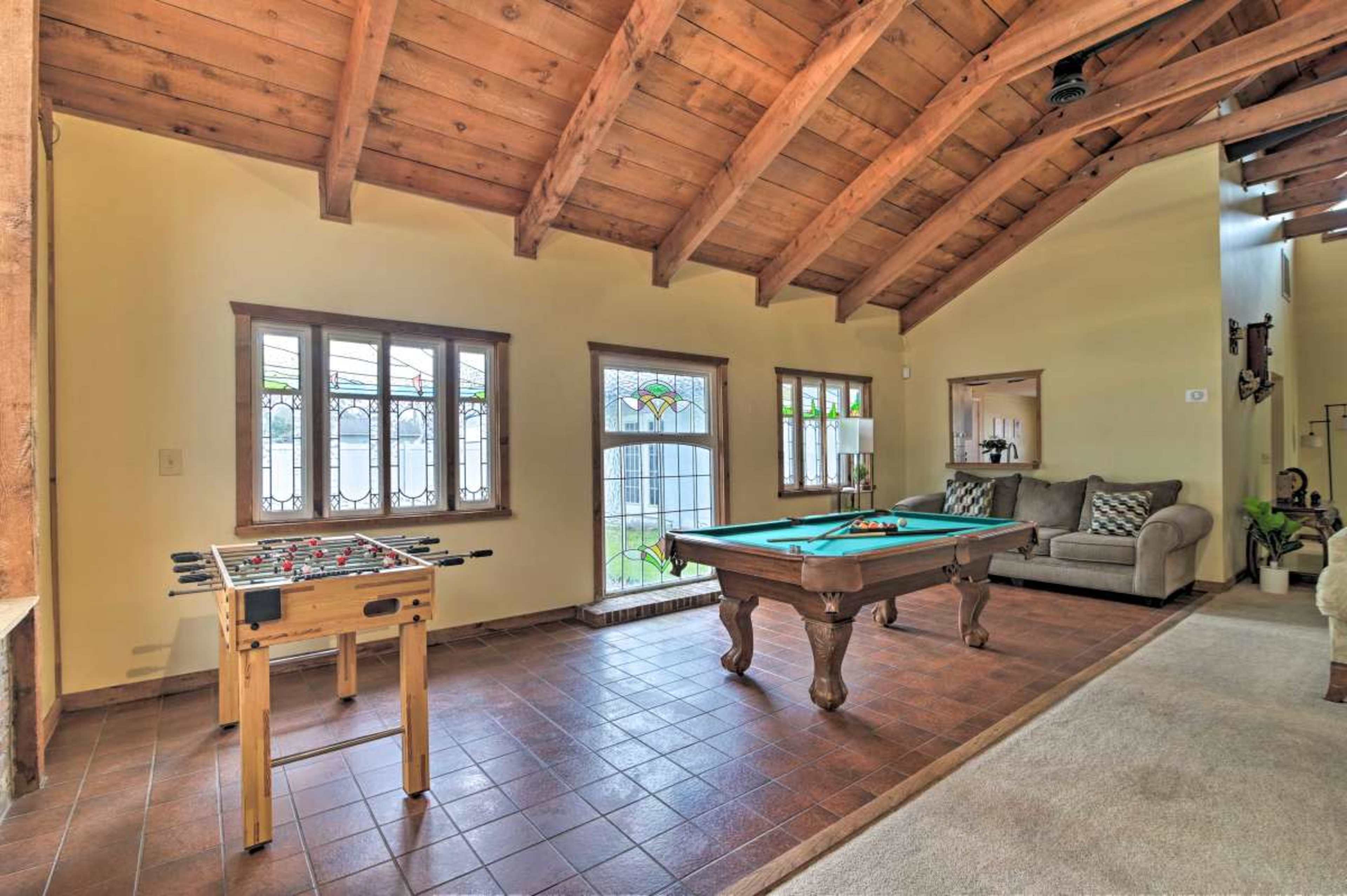 A cozy game room featuring a foosball table and a billiard table, with large windows and a wooden beam ceiling.