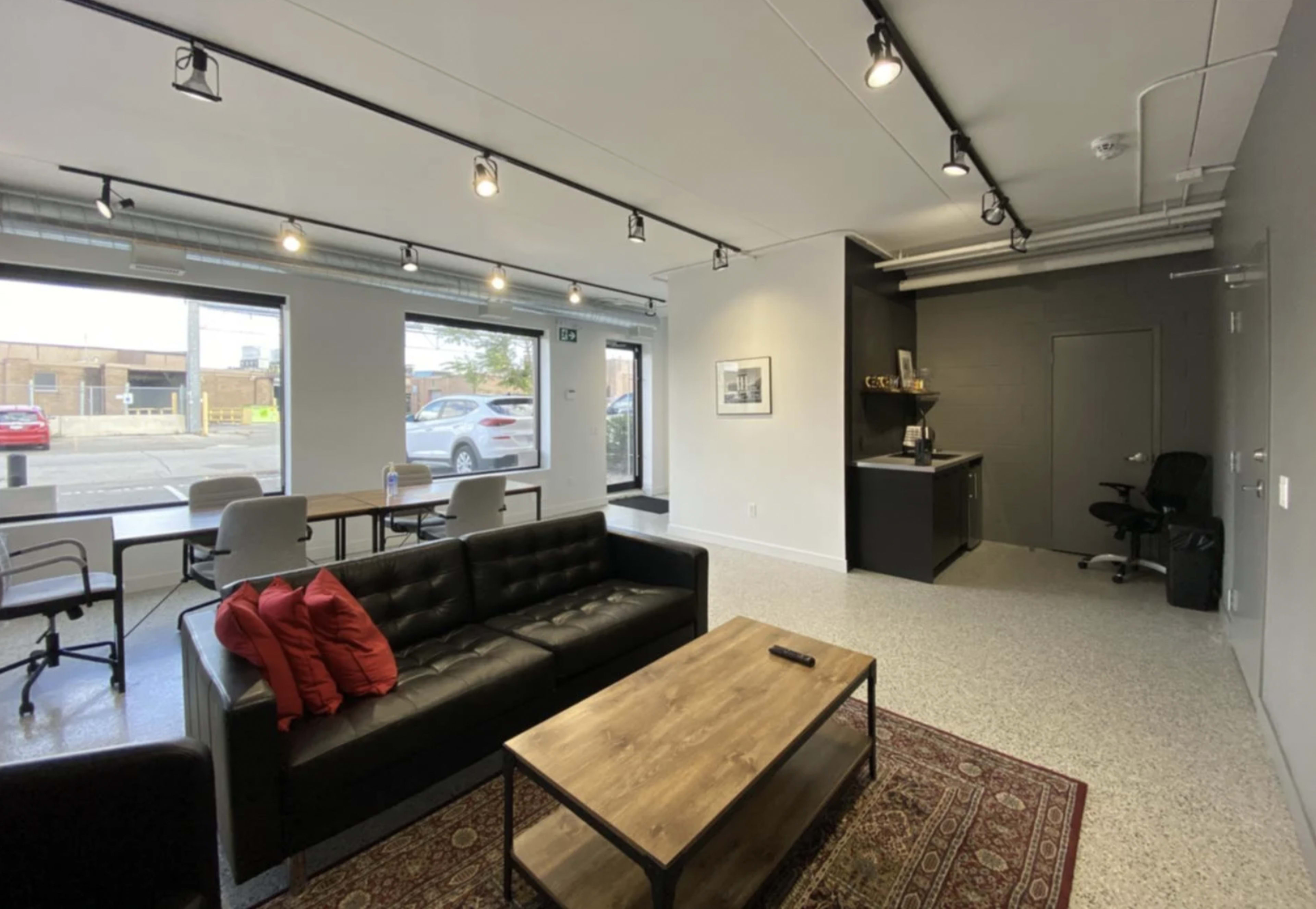 A modern, open lounge area with a black couch, a wooden coffee table, and a kitchenette in the corner, featuring large windows allowing natural light to enter.