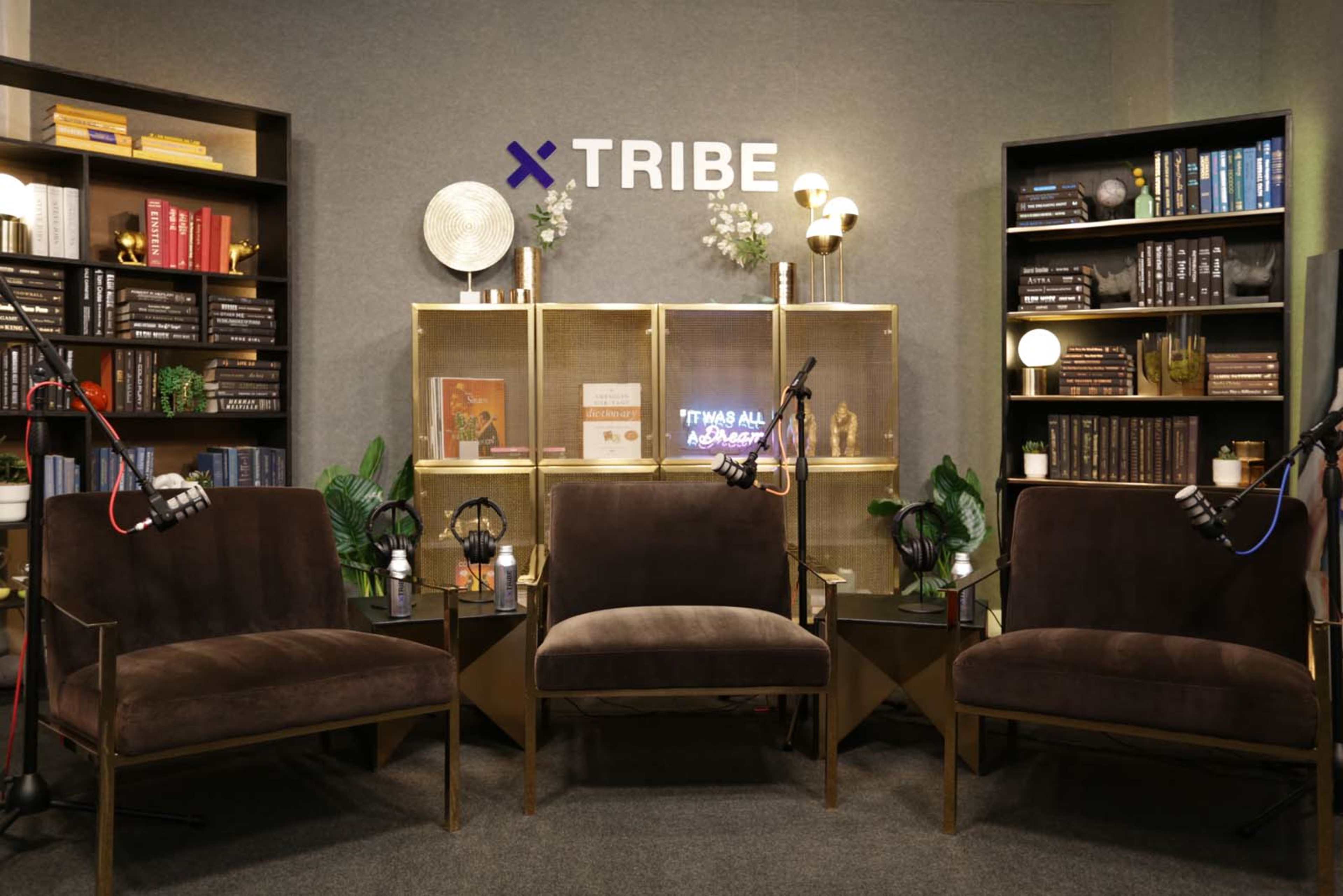 A neatly arranged set with three brown chairs, a decorative background featuring shelves with books, plants, and a sign that reads "TRIBE."