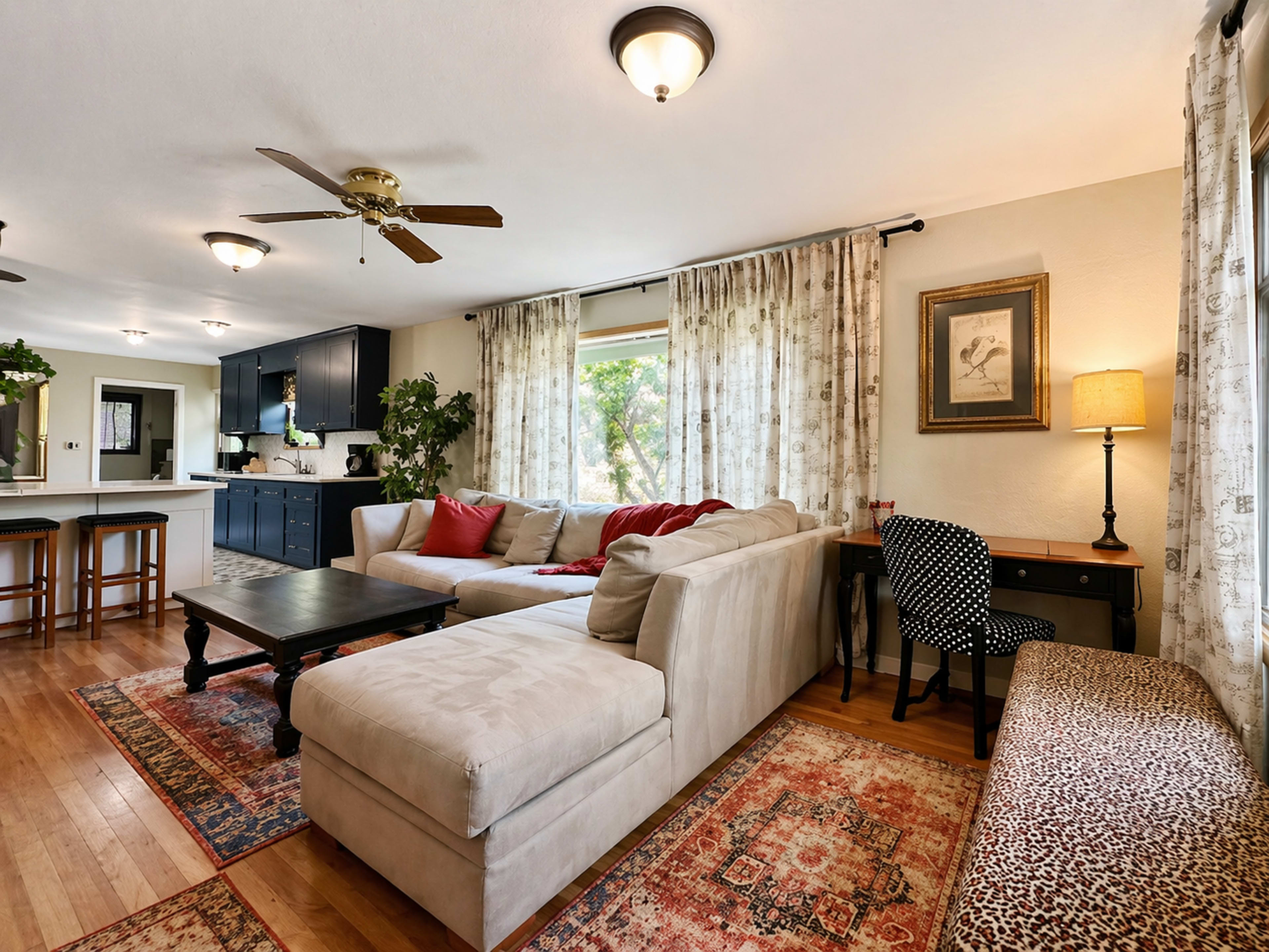 The image shows a cozy living room with a sectional sofa, a coffee table, a plant, and a desk beside a window, featuring warm lighting and decorative rugs.