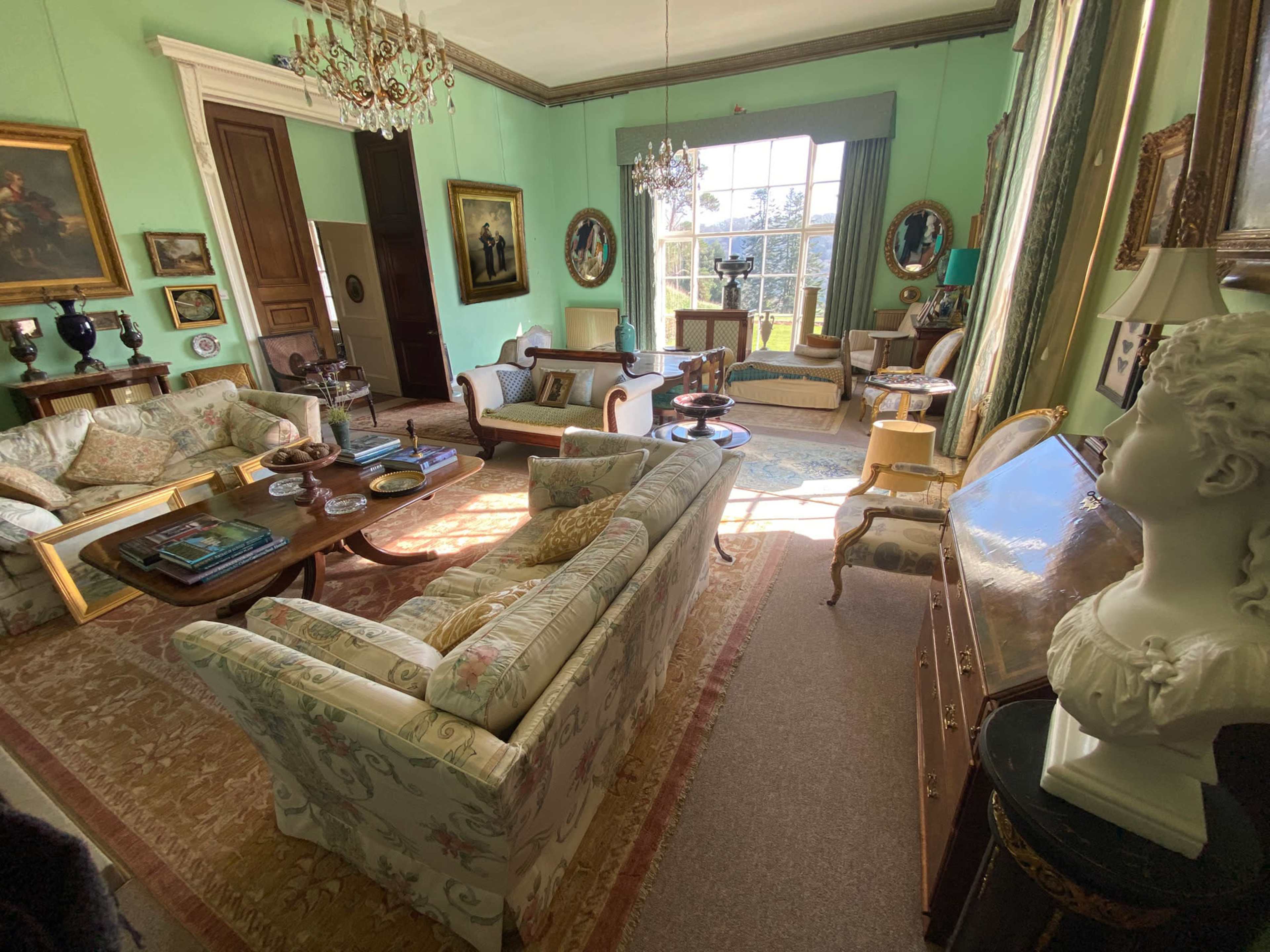 The room features elegant furniture, including sofas and a wooden table, with large windows allowing natural light to illuminate the space filled with art and decorative items.