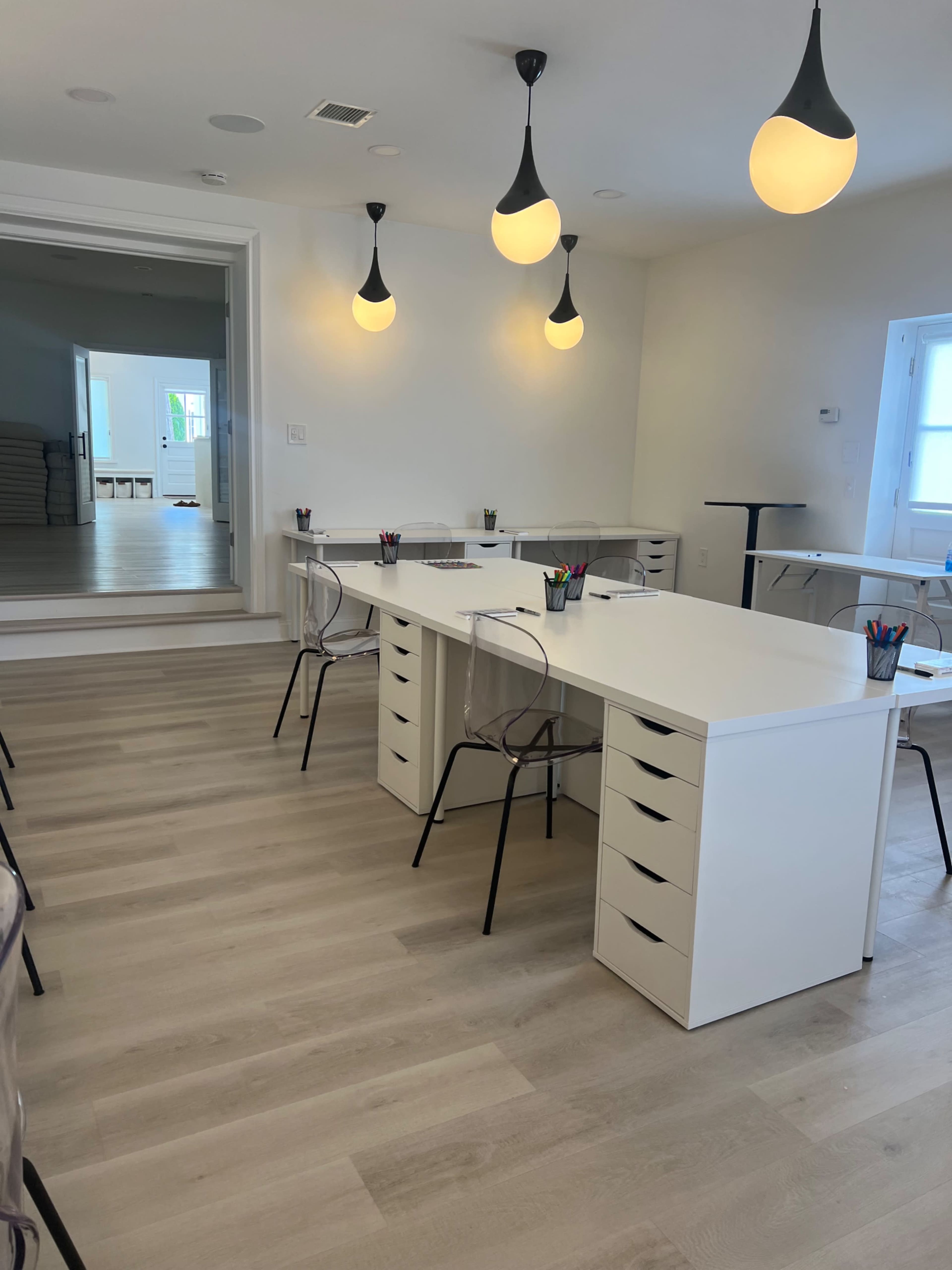 A bright, minimalist office space featuring multiple white desks with transparent chairs and pendant lighting.