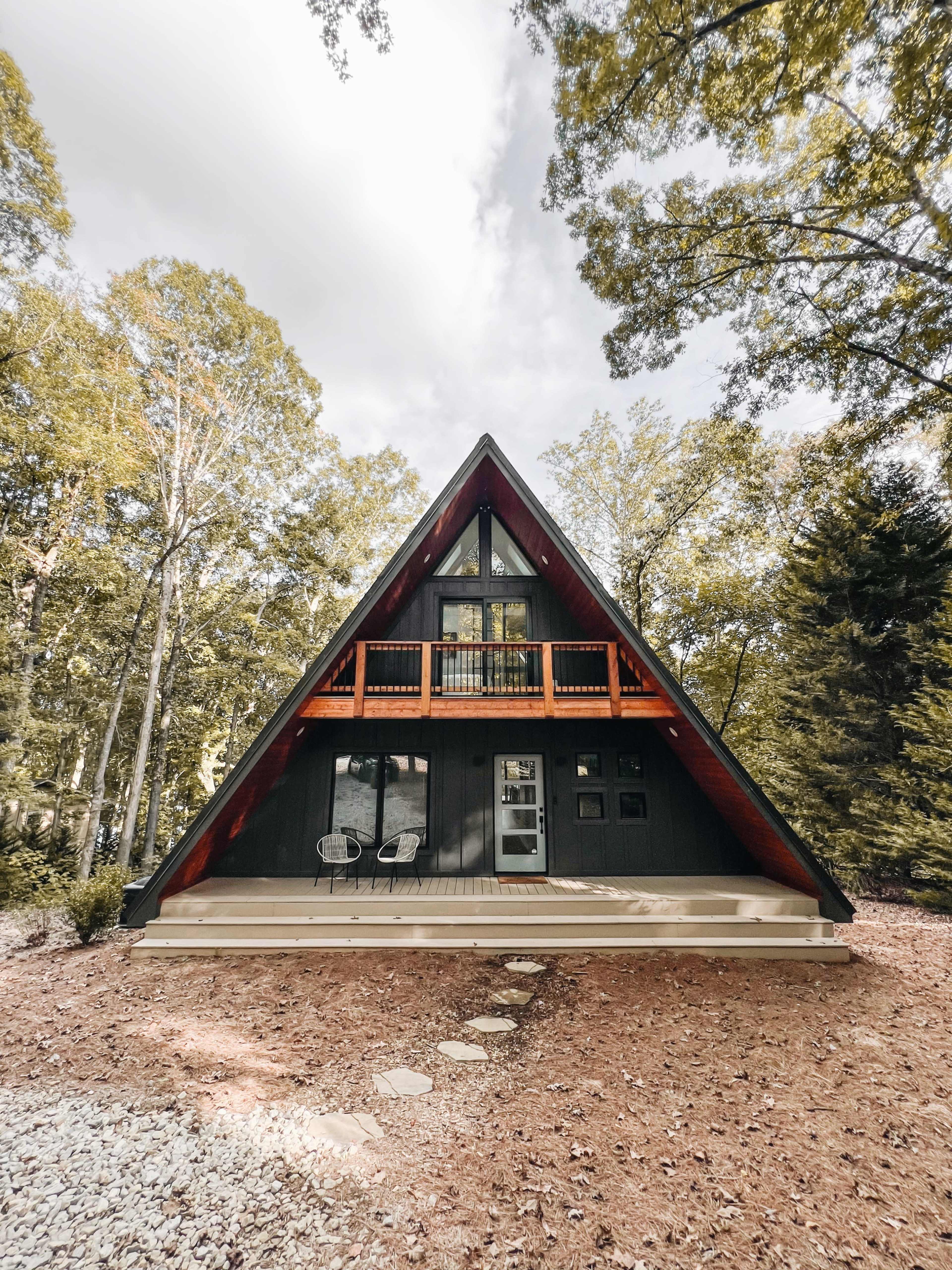 A triangular, A-frame cabin surrounded by trees, with a front porch and stone pathway leading to the entrance.