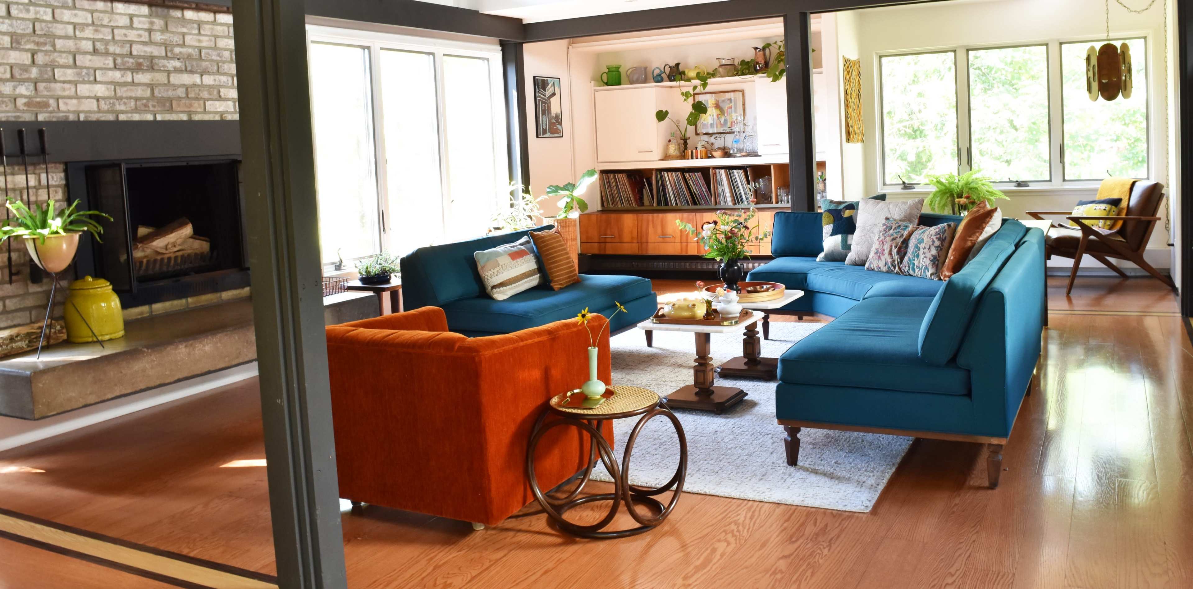 The image shows a living room with a blue sectional sofa, an orange chair, a wooden coffee table, and large windows allowing natural light.