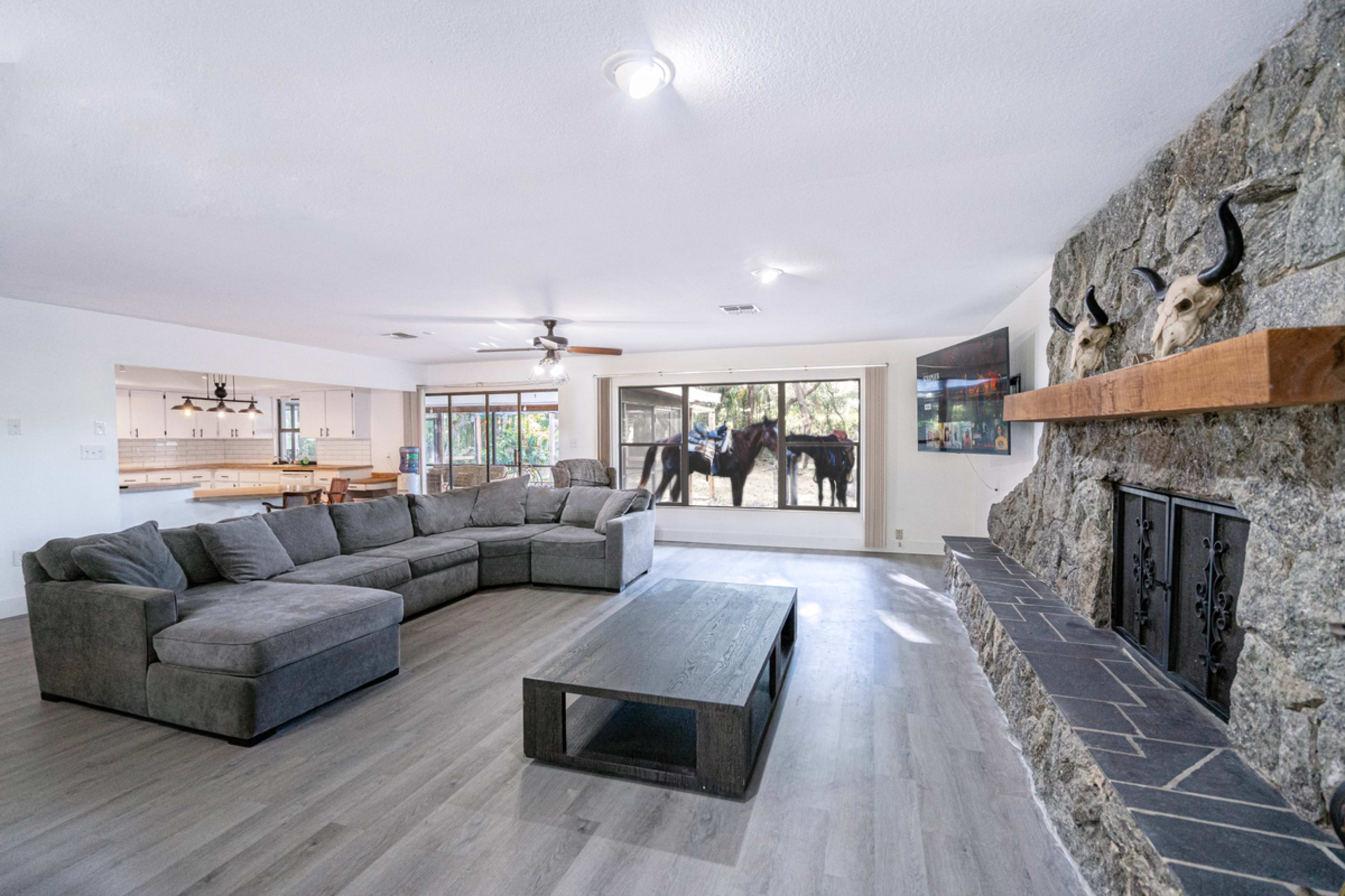 A spacious living room features a large sectional couch and a stone fireplace, with a view of horses outside through large windows.