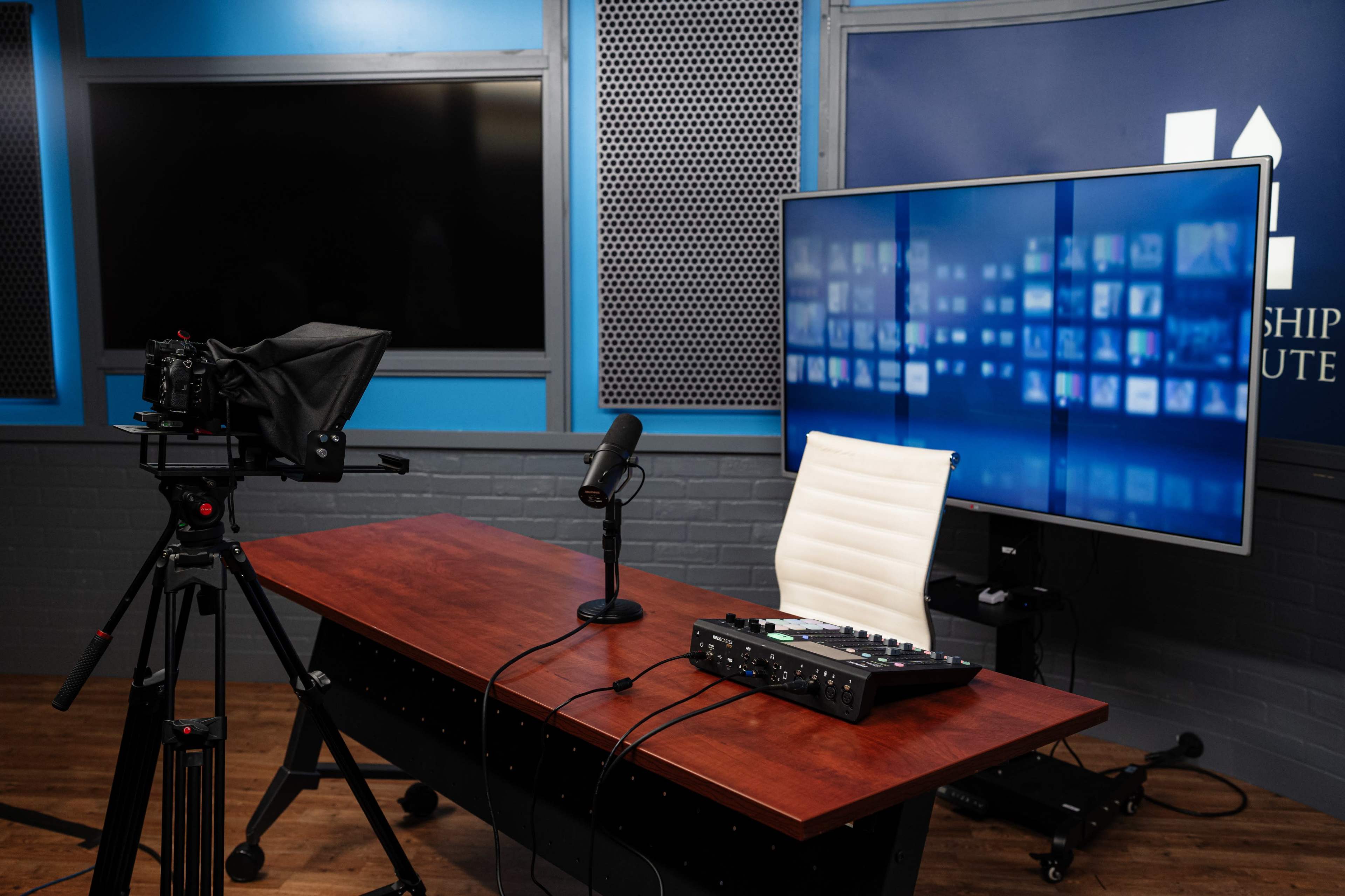 A recording studio setup features a wooden table with a microphone, a control panel, a white chair, a camera on a tripod, and a large screen displaying multiple images in the background.