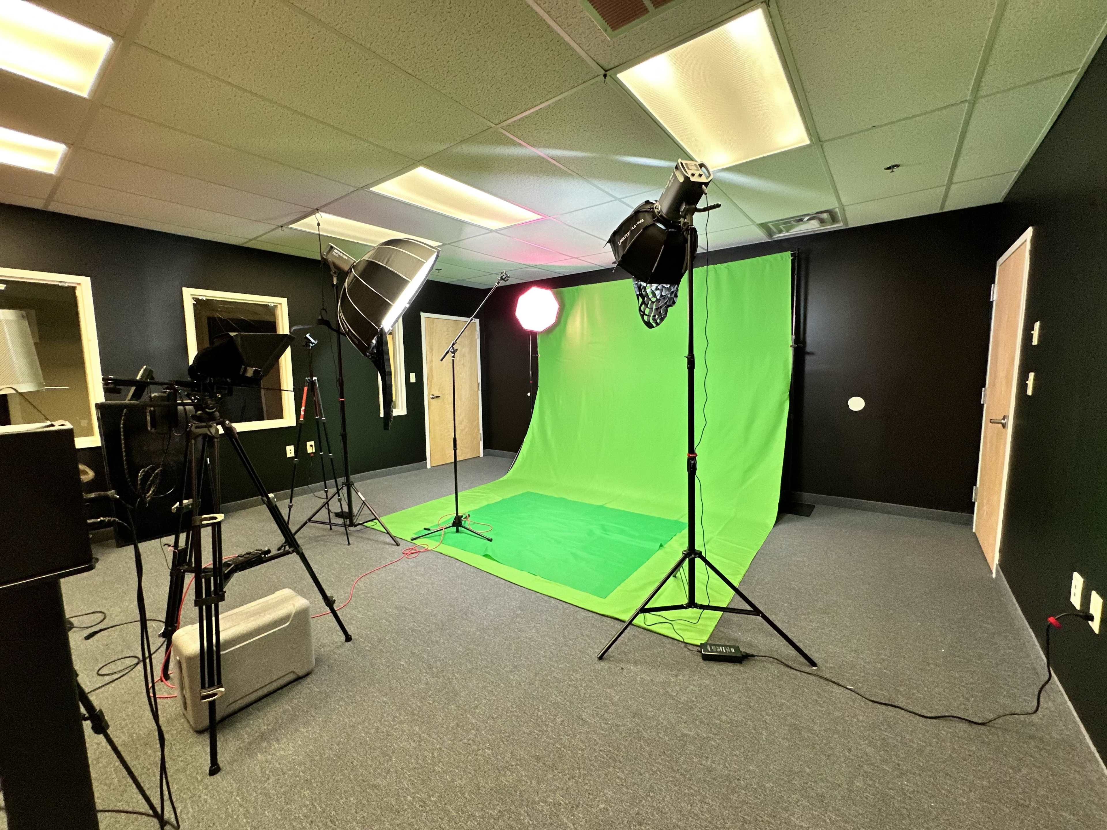 The image shows a studio setup with a green backdrop, lighting equipment, and cameras in a room with gray carpet and black walls.