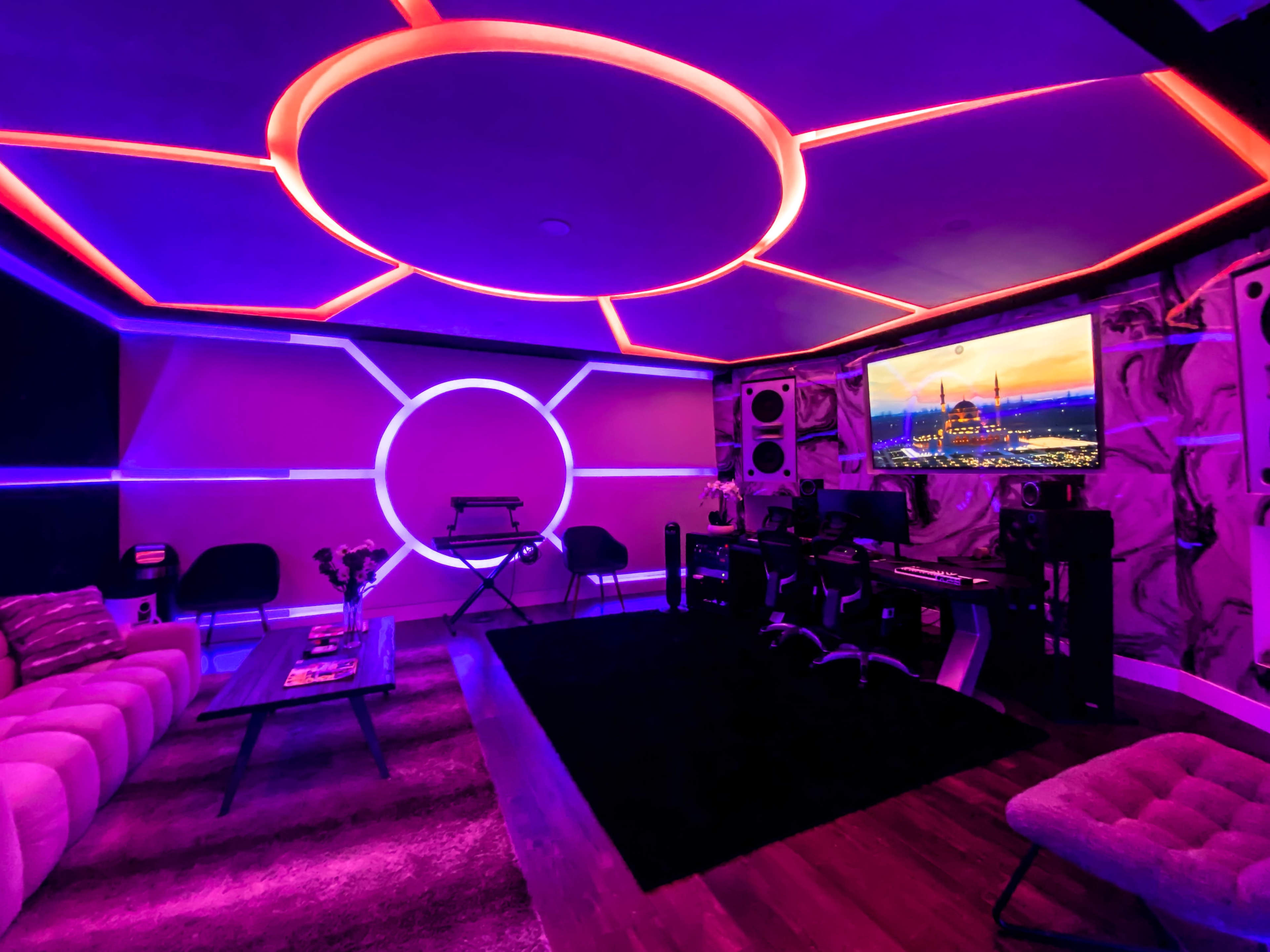 The image shows a modern room featuring a purple and pink LED lighting design, a large screen displaying a cityscape, and a workspace with computer equipment.