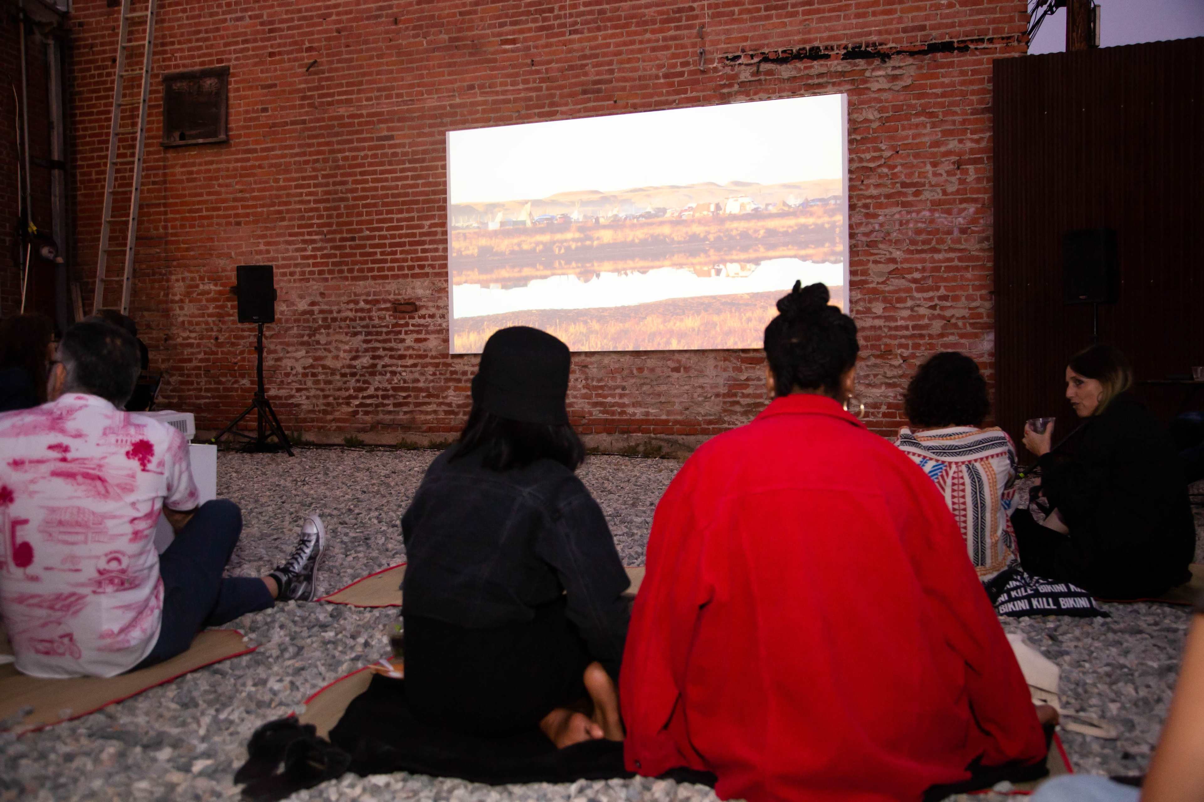 A group of people sits on the ground watching a film projected onto a brick wall outdoors.