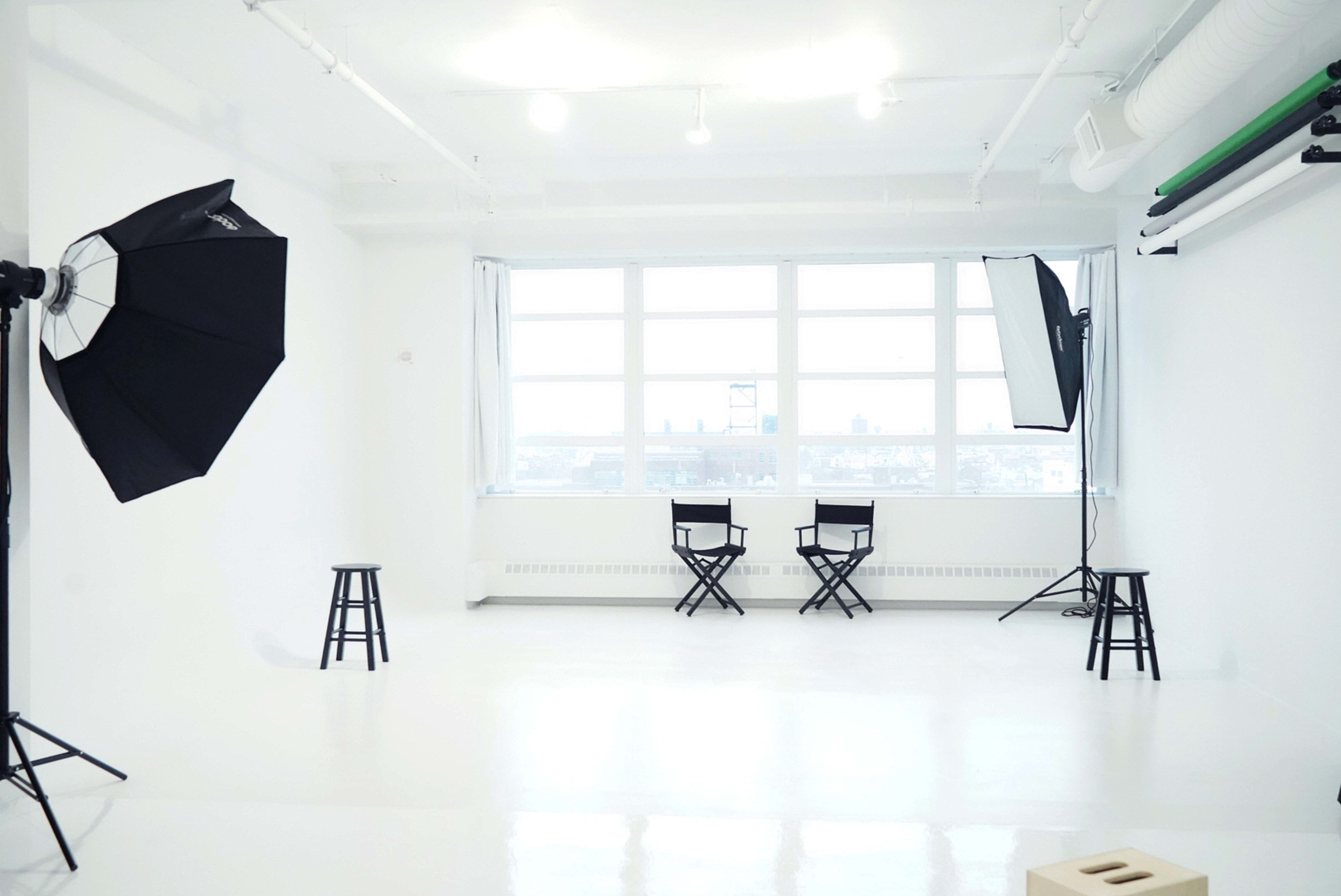 A bright, empty studio with two director's chairs and photography lighting equipment set up near large windows.