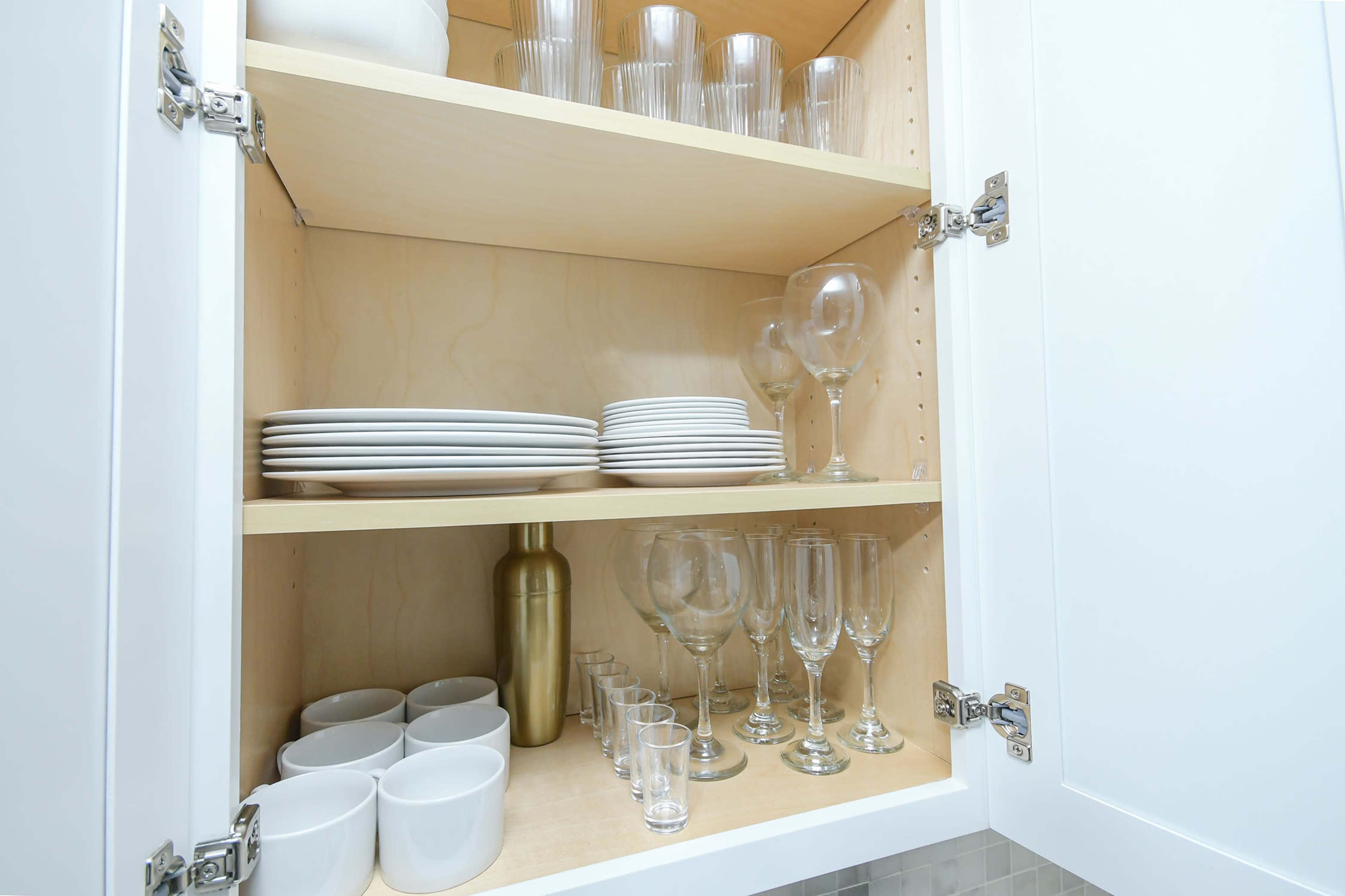 The image shows a kitchen cabinet interior with neatly arranged white plates, glassware, and cups on wooden shelves.