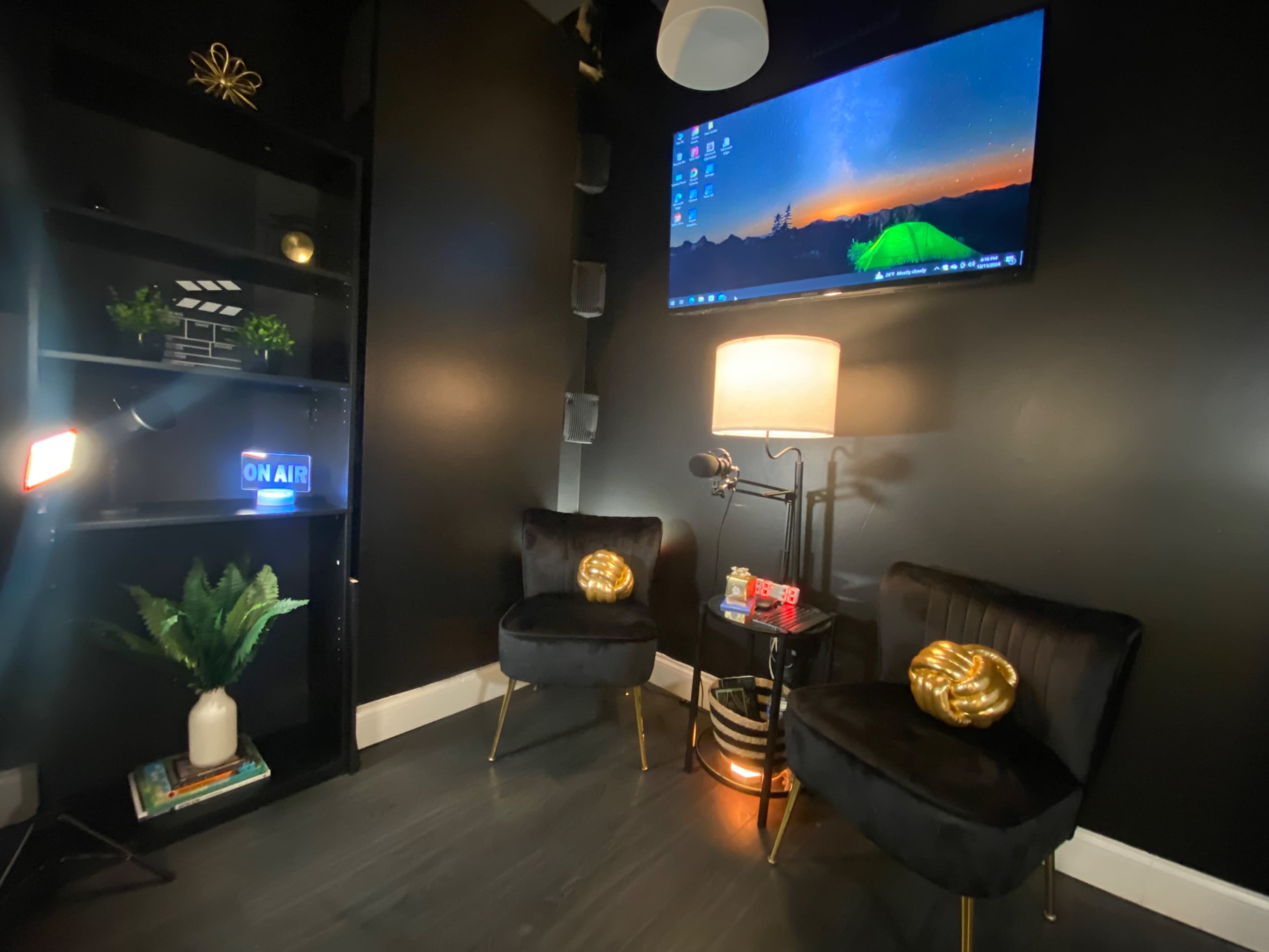 The image shows a small, modern room with two black velvet chairs, a lamp, a microphone, a wall-mounted TV displaying a landscape, and a shelf with decorative items.