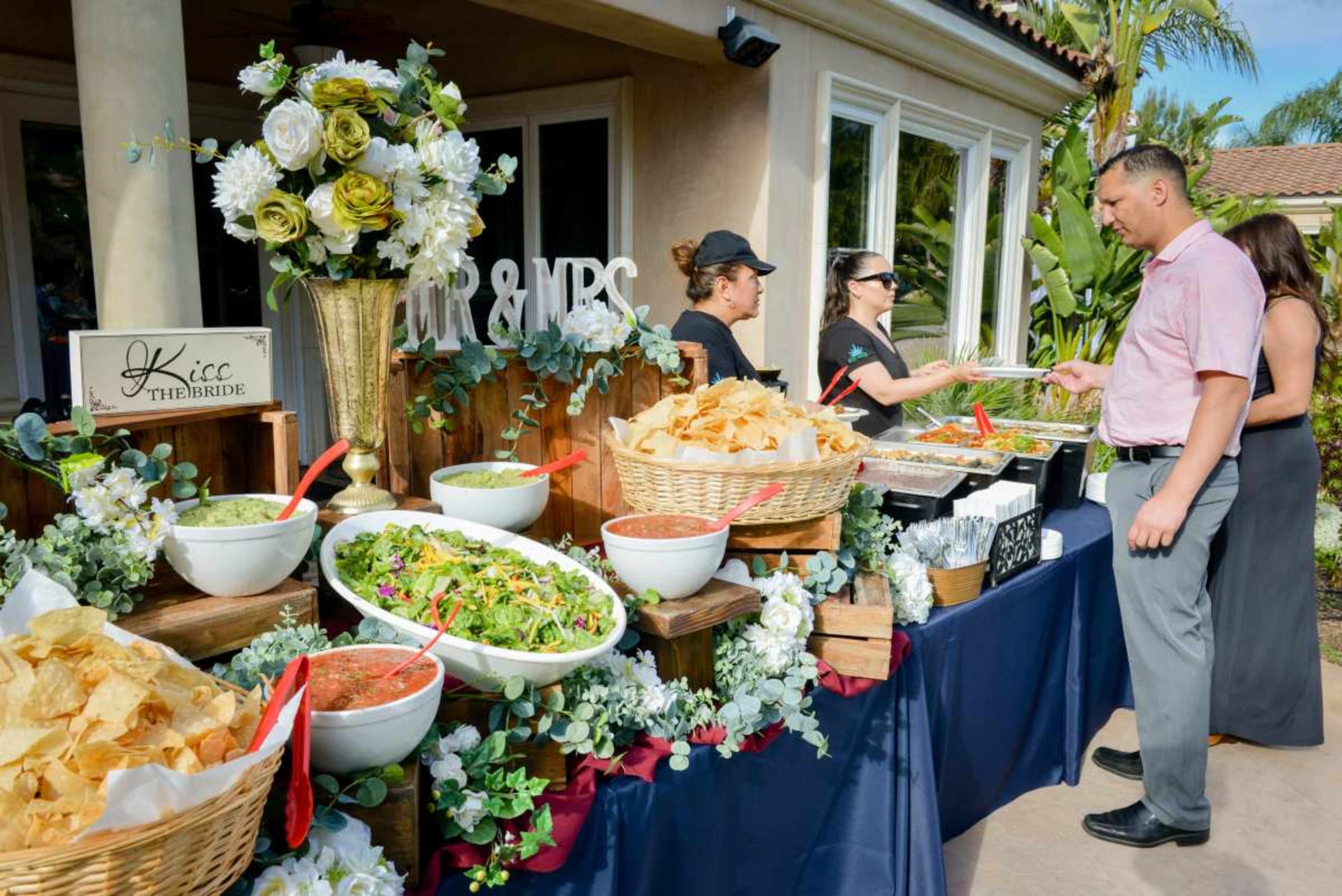 A catering table displays a variety of food options, including chips, salads, and salsas, while guests engage with staff in an outdoor setting.