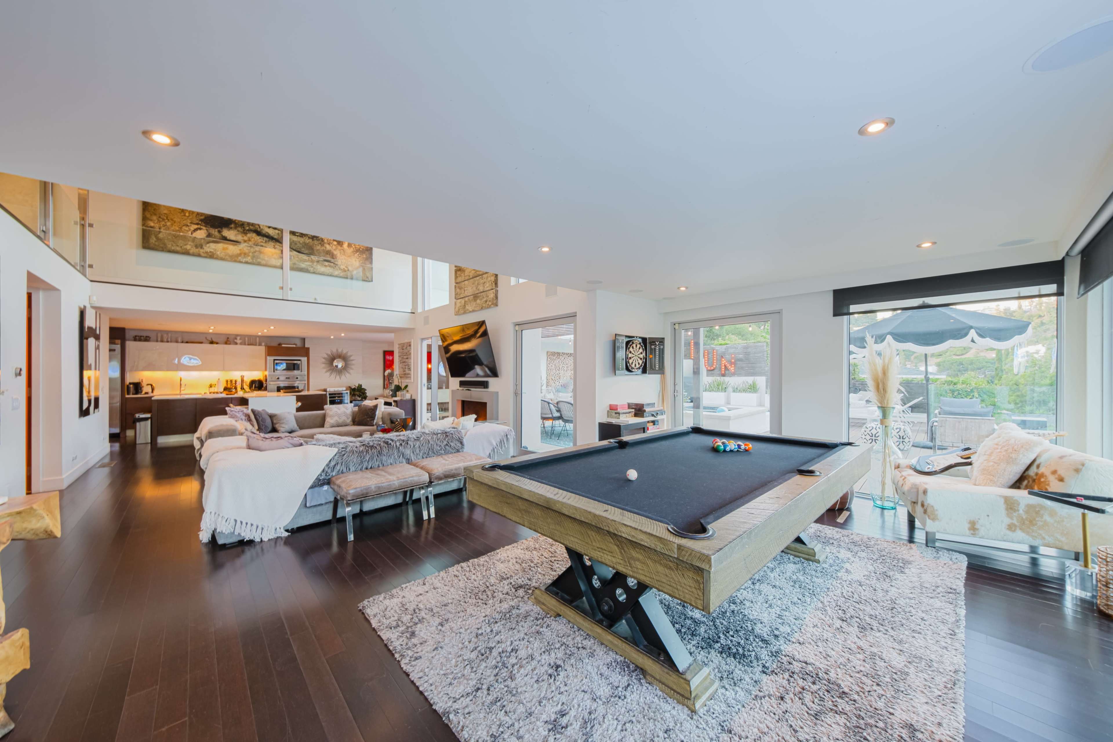 The image shows a modern living space featuring a pool table in the center, surrounded by a cozy seating area and large windows that open to an outdoor patio.
