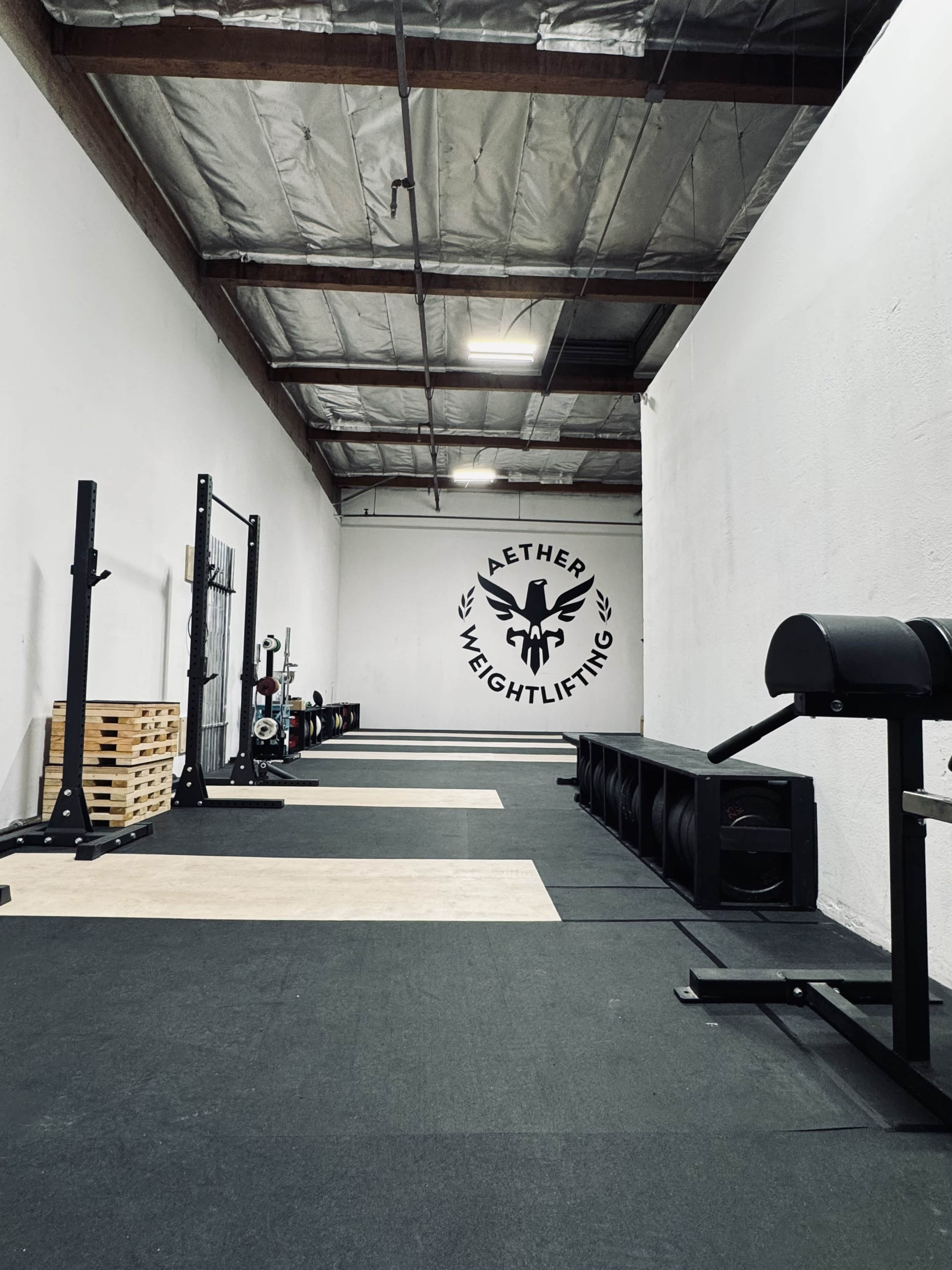 The image shows a spacious weightlifting gym with black flooring, weightlifting equipment, and a large logo on the wall.