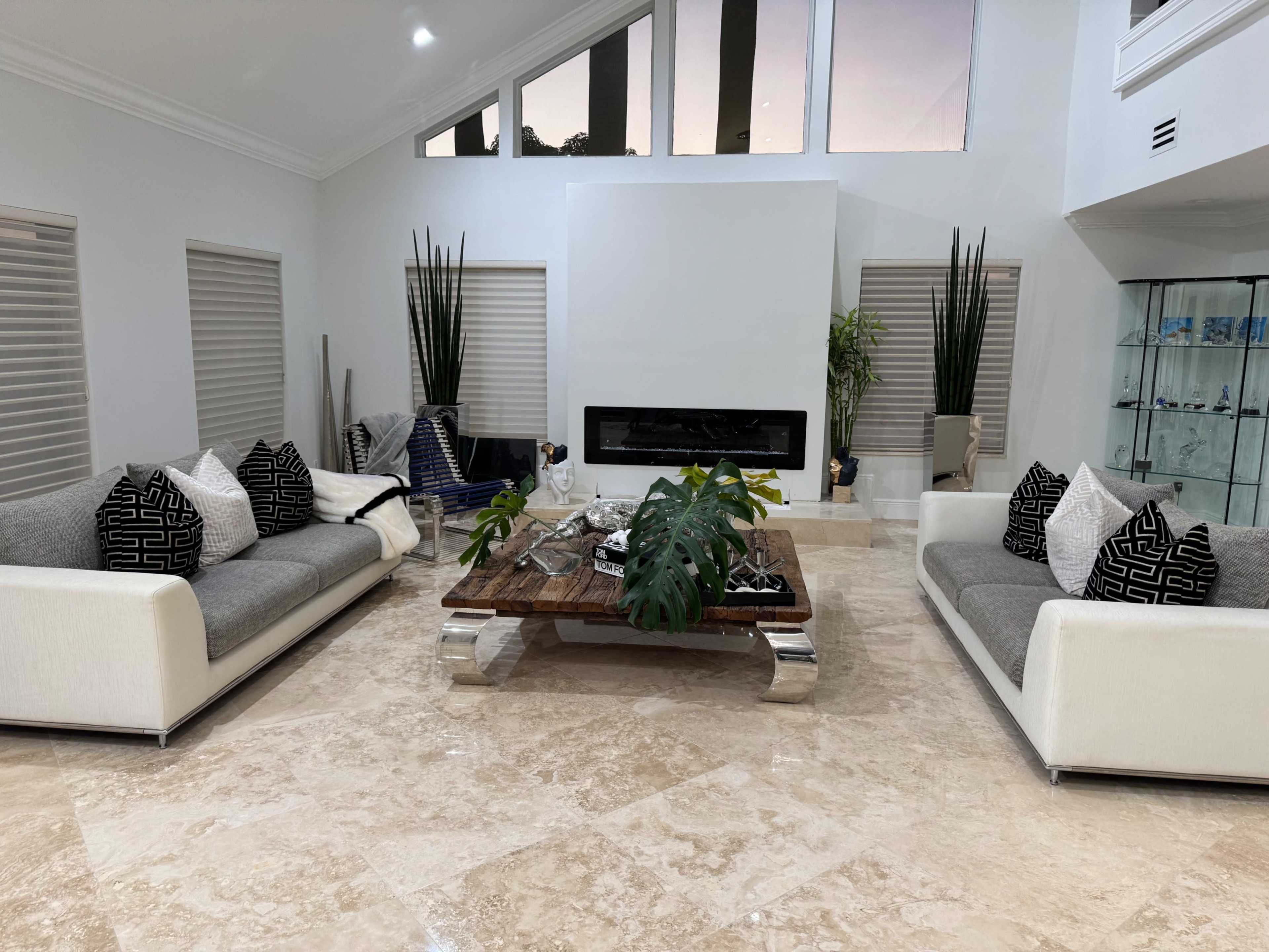 The image shows a modern living room with two sofas, a large coffee table at the center, and a fireplace, all surrounded by large windows and decorative plants.